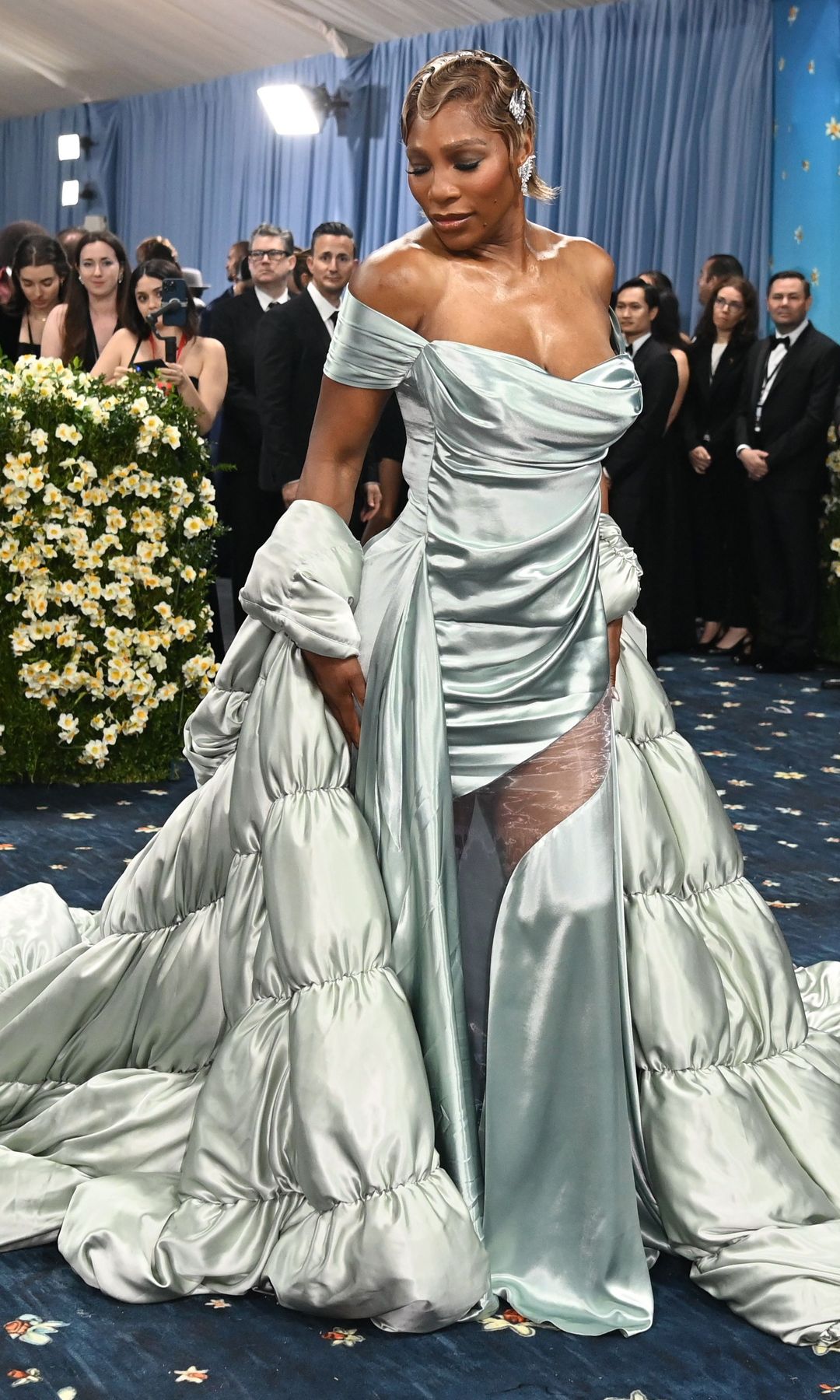 Serena Williams wears a very pale blue off the shoulder mini dress with matching quilted cape. Her hair is styled in vintage finger waves.