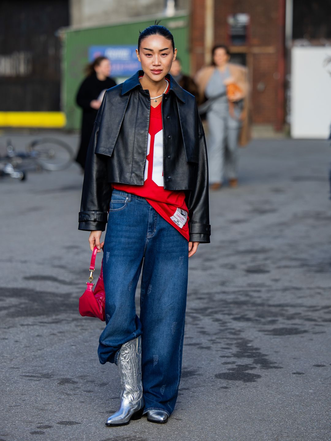 A guest wears black leather jacket, denim jeans, silver cowboy boots, red bag outside Gestuz during the Copenhagen Fashion Week 