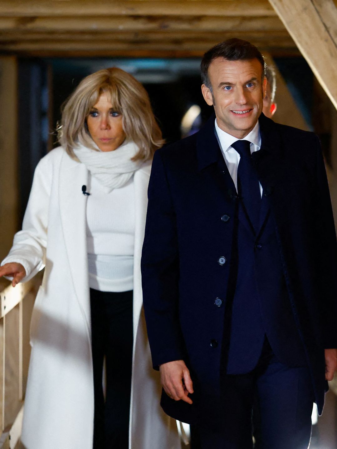 Brigitte and Emmanuel Macron walk along a wooden bridge inside a dimly lit structure. She wears a white coat and scarf, while he’s in a navy suit and coat.