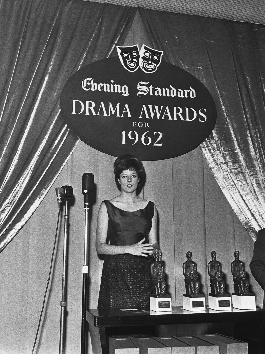 Maggie Smith is standing at a podium, accepting an award at the Evening Standard Drama Awards in 1962. She wears a sleeveless black dress and has short hair. The backdrop features dramatic curtains and large awards displayed behind her. Her posture is composed, and she looks focused, exuding an air of professionalism and confidence as she delivers her acceptance speech.