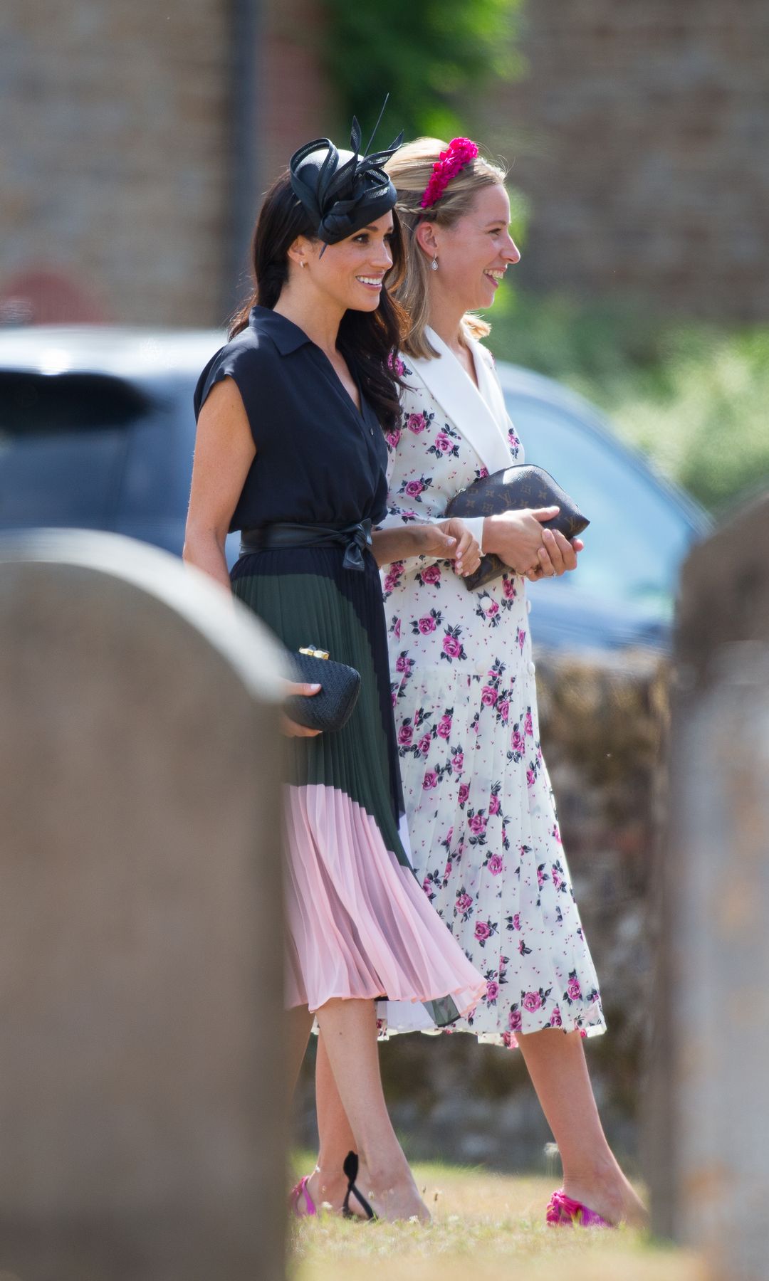 Meghan Markle walks through the church yard with another wedding gest who wears a white and pink floral midi dress with matching headband