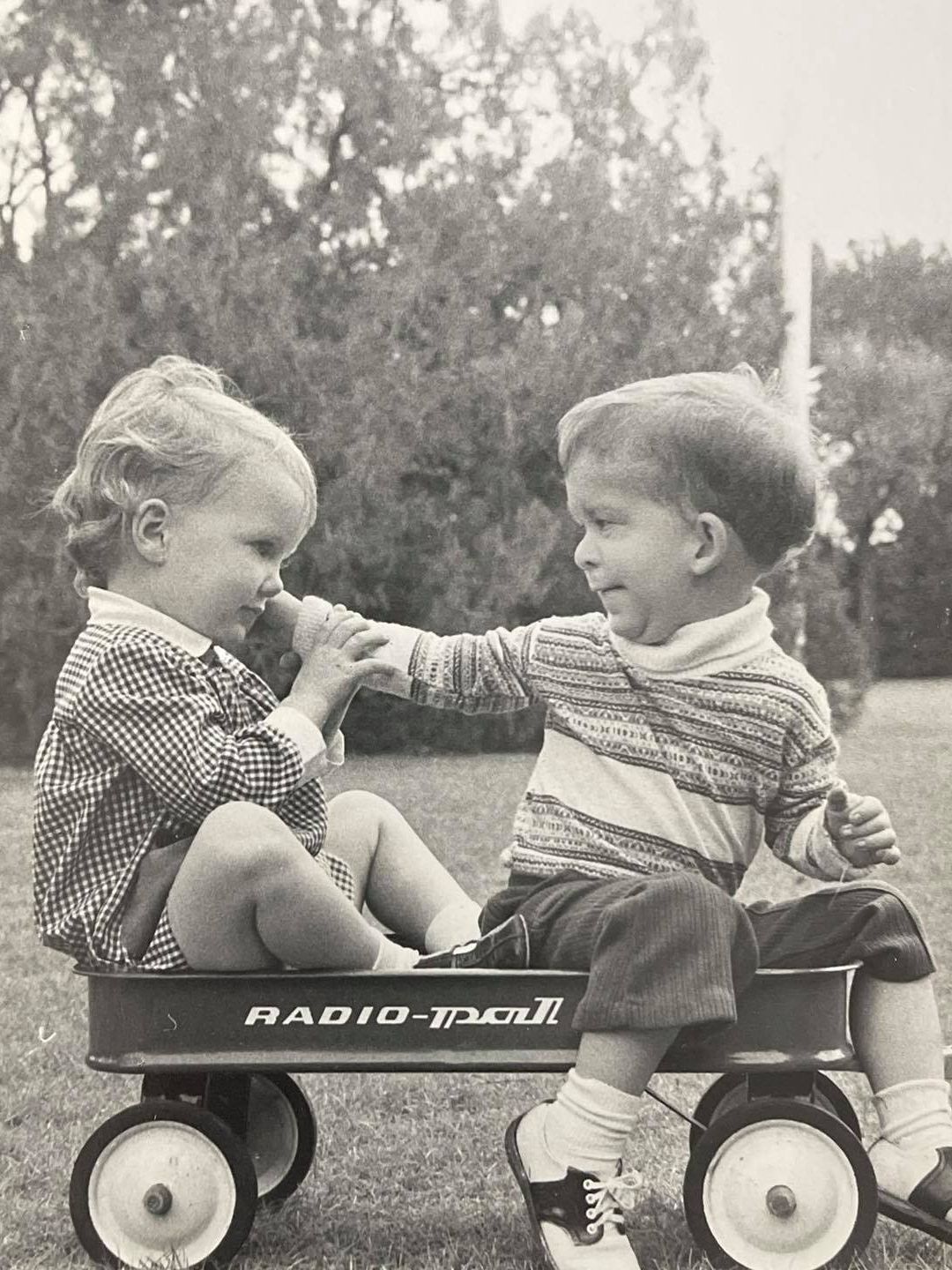 Black and white photo of two young children sat in a cart, Ree is on the left and her brother is on the right, reaching out to touch her cheek