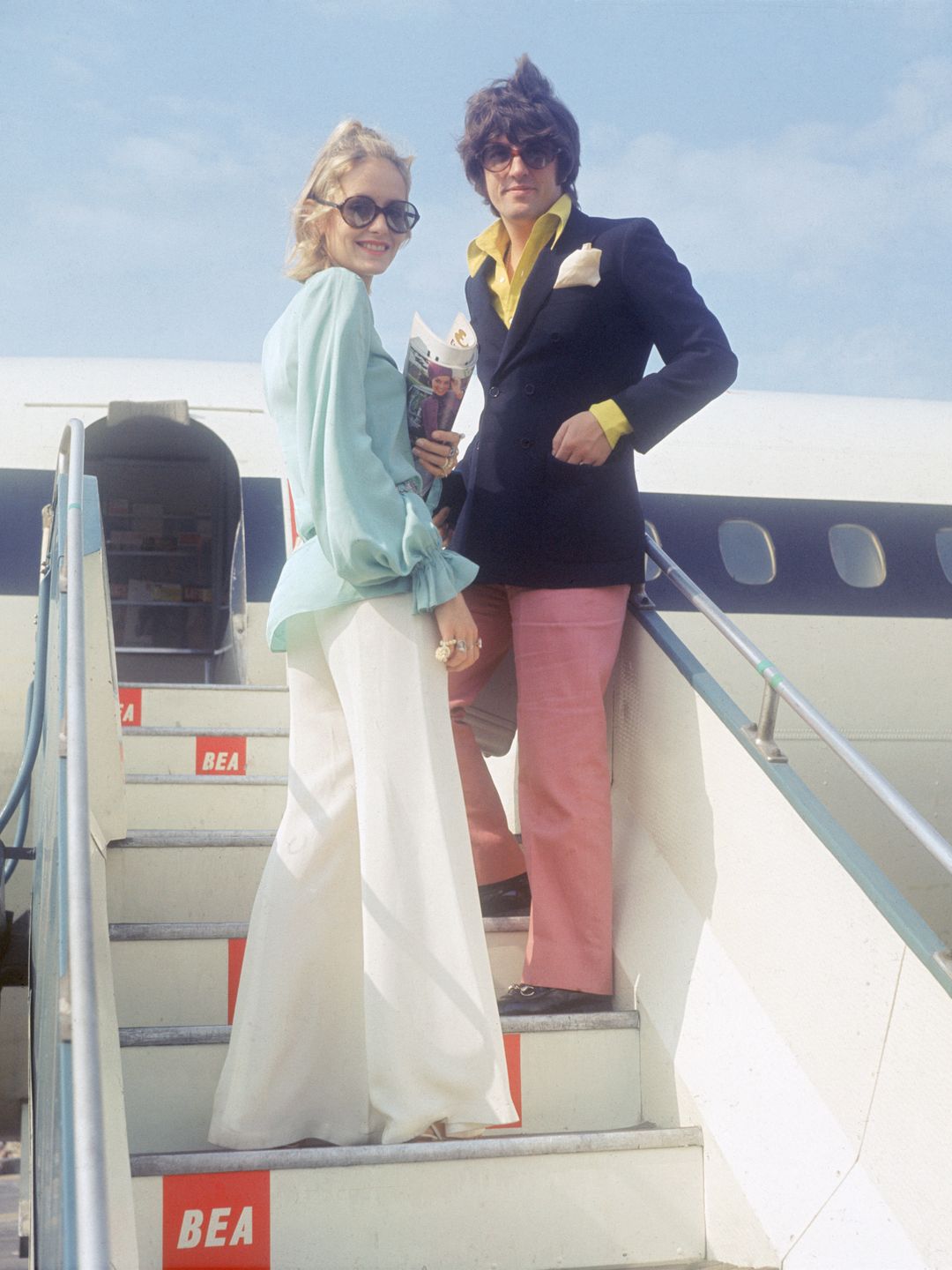 Chegando ao aeroporto com estilo, Twiggy e seu parceiro fazem uma entrada ousada. Ela ostenta calças largas, tons pastéis e óculos de sol grandes, um visual clássico para fashionistas jet-set da época. Este estilo polido, mas relaxado, sinaliza a mudança para uma moda mais glamourosa e amigável para viagens.