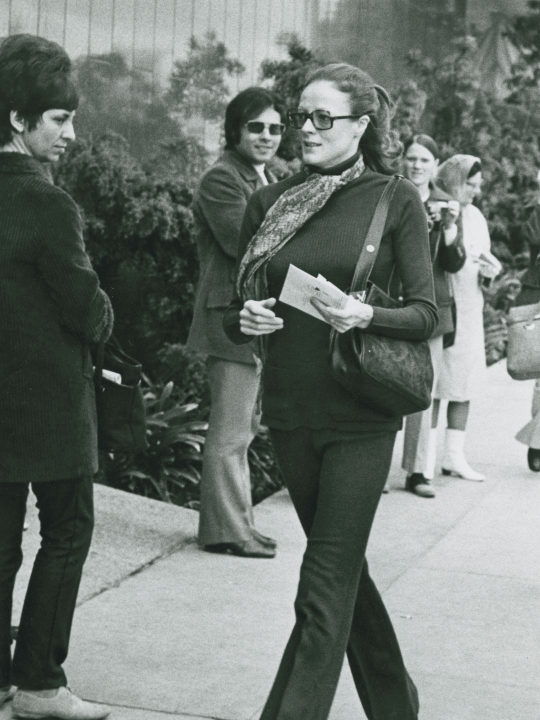 Maggie Smith is captured walking down a street in the 1970s, wearing a form-fitting turtleneck, flared pants, and large glasses. Her hair is pulled back, and she carries a bag over her shoulder. She holds papers in her hand, moving confidently through a crowd. The scene is casual yet stylish, showcasing her natural elegance and timeless fashion sense from the era.