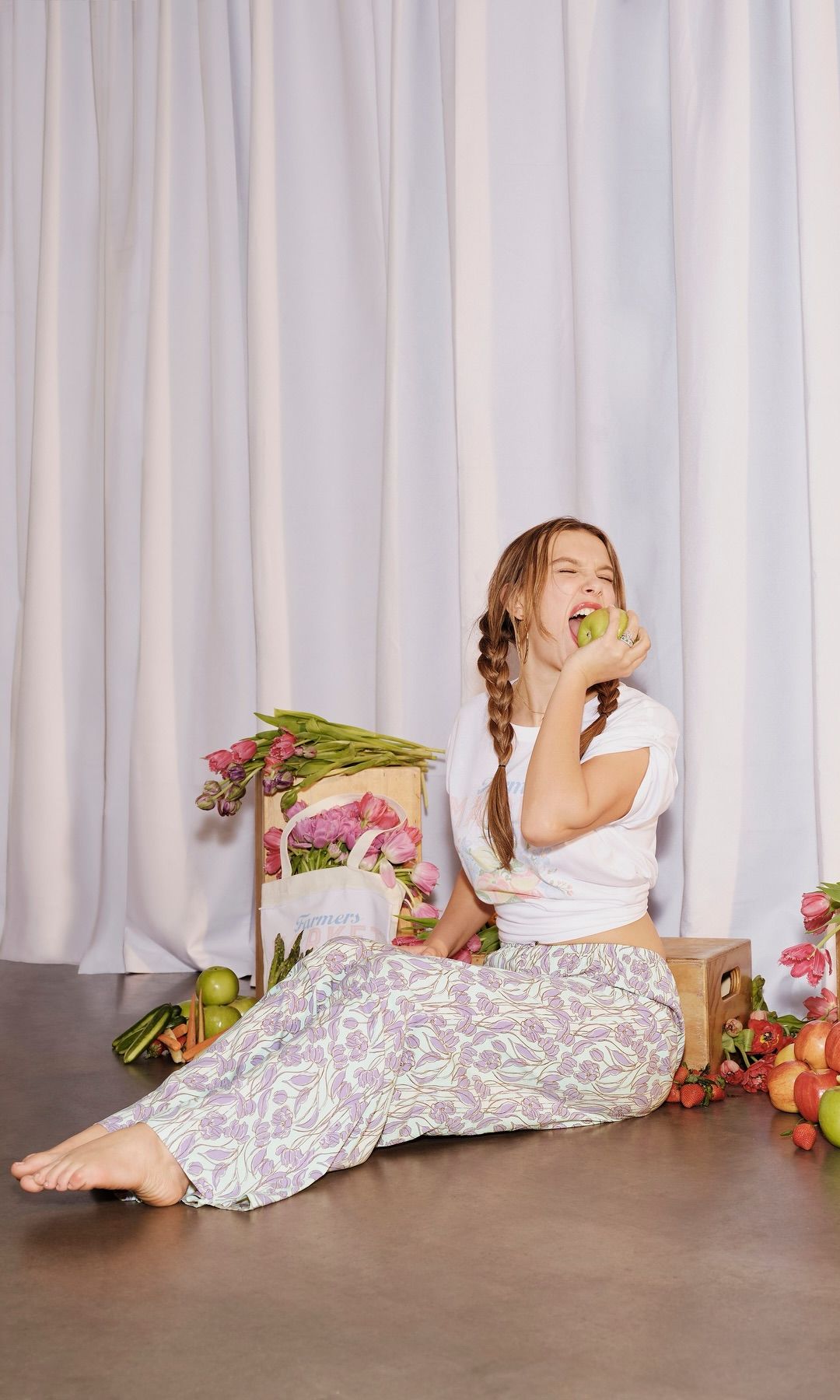 Millie Bobby Brown poses on a crate alongside fresh fruit and vegetables wearing a t-shirt and floral pants
