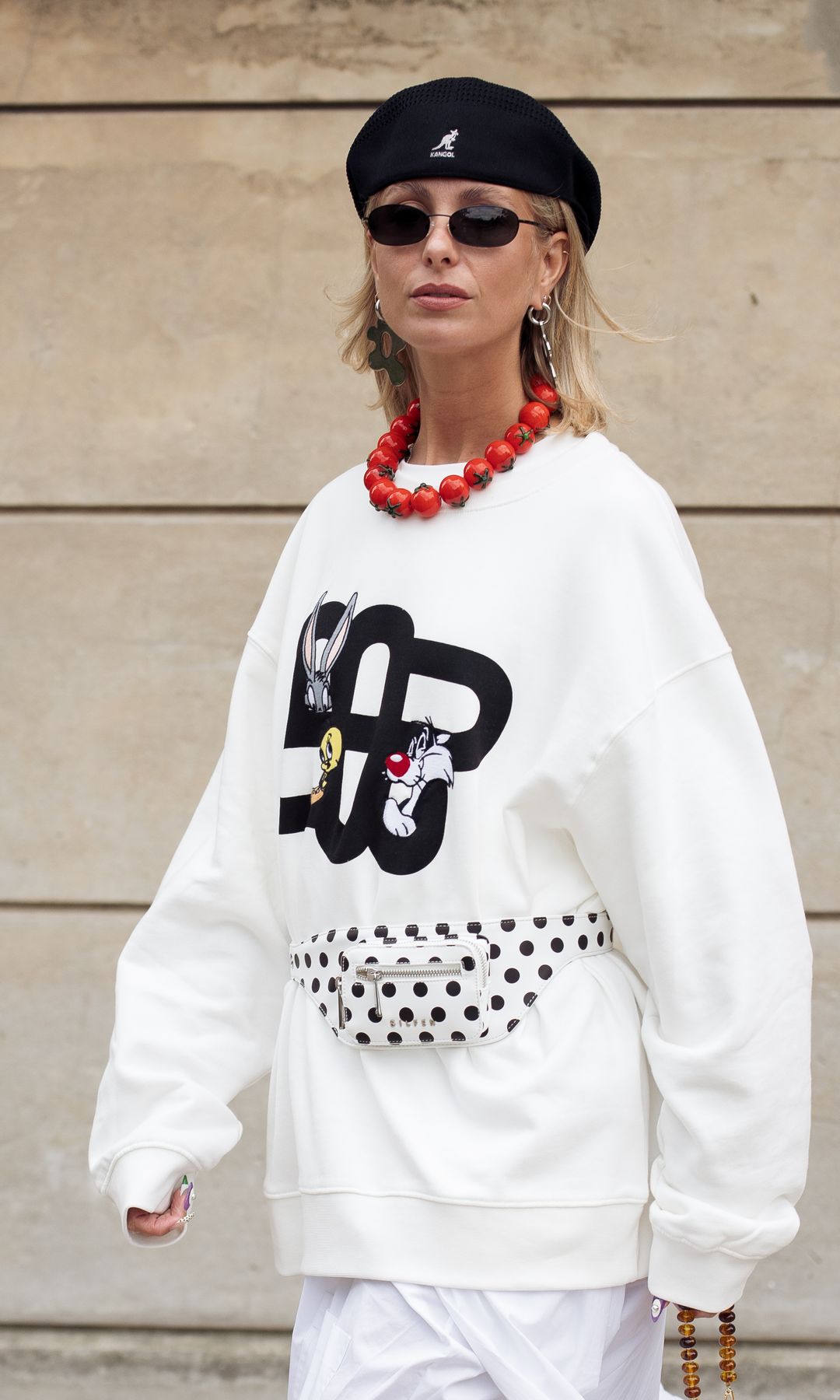 A guest wears white skirt, white sweater with animation heroes from Munthe and Looney Tunes collaboration, red shoes and black hat outside Munthe show during Copenhagen Fashion Week 