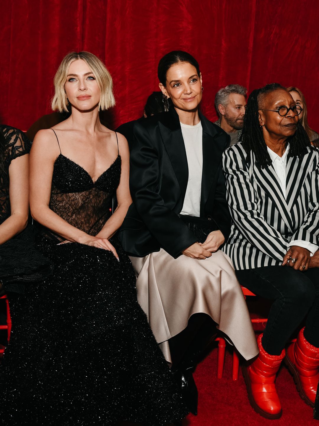 The image depicts a group of three women seated in the front row at a fashion event, against a vibrant red velvet backdrop. They are dressed in glamorous and diverse outfits, representing a mix of high-fashion elegance and personal style. From left to right: The first woman dons a black, sparkling gown with thin straps and sheer elements, creating a balance of glamour and allure. Her short, blonde bob haircut frames her face beautifully. The second woman is Katie Holmes, wearing her previously described outfit. She pairs an oversized black blazer with a white t-shirt and a flowing champagne satin skirt. Her hair is slicked back neatly, and her overall style exudes sophisticated minimalism. The third woman, Whoopi Goldberg, wears a striking black-and-white striped blazer over a white shirt, paired with black pants. She adds a playful touch with bold red boots. Her signature glasses and dreadlocks complete her statement look. The group sits on red chairs, and their outfits stand out against the red backdrop. The image captures a moment of camaraderie and style, showcasing the unique fashion choices of each woman while celebrating diversity and individuality.