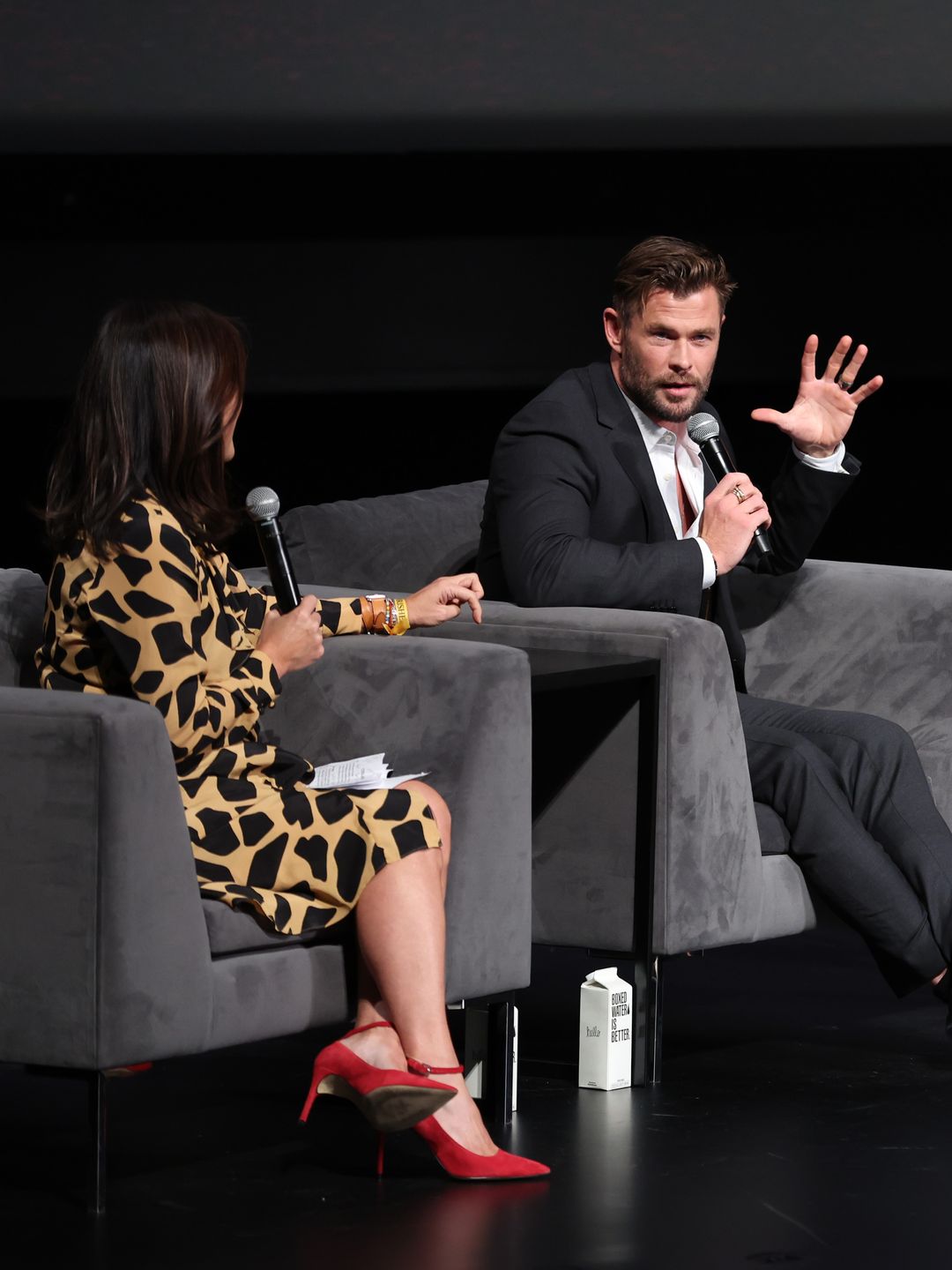 Chris Hemsworth sat on a stage being interviewed by another person, who has her back to the camera