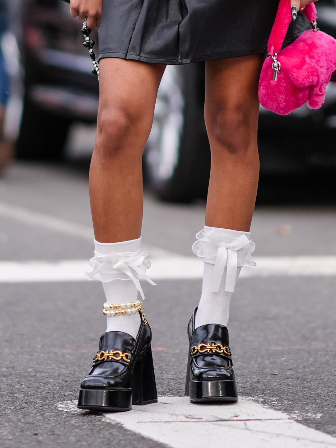 A guest wears sunglasses, earrings, a white shirt, a black mini leather skirt with bow details, a pink fluffy Coach bag, black leather platform shoes, white socks outside Coach, during New York Fashion Week