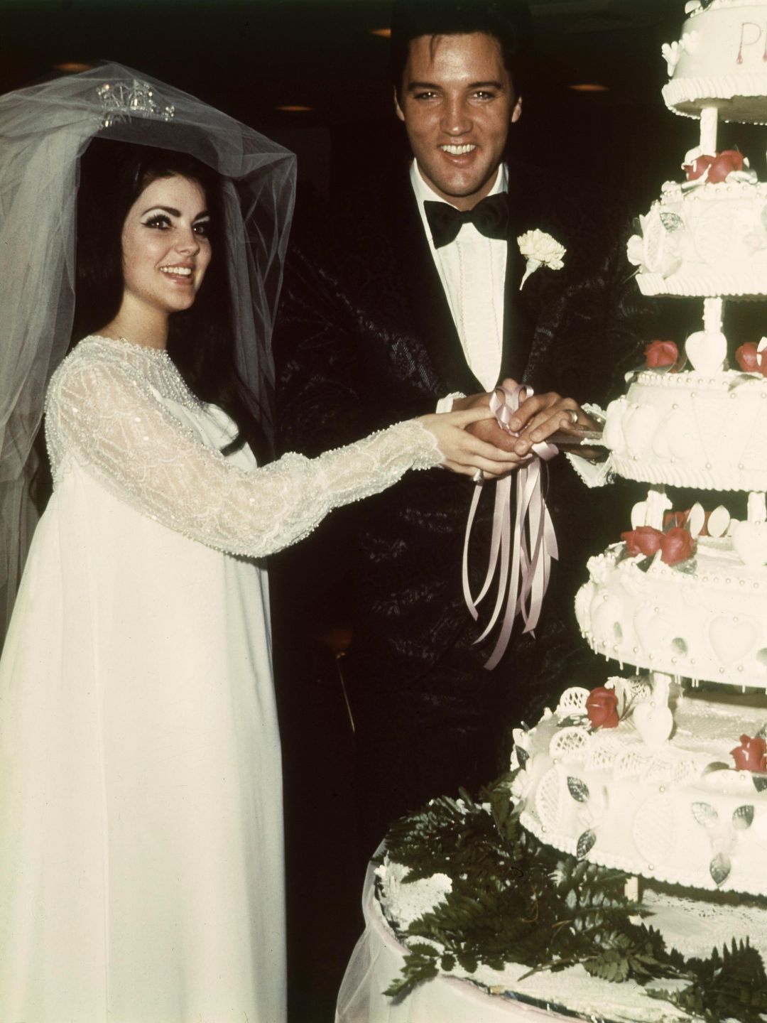 Priscilla Presley wearing a floaty wedding dress while cutting her wedding cake with Elvis