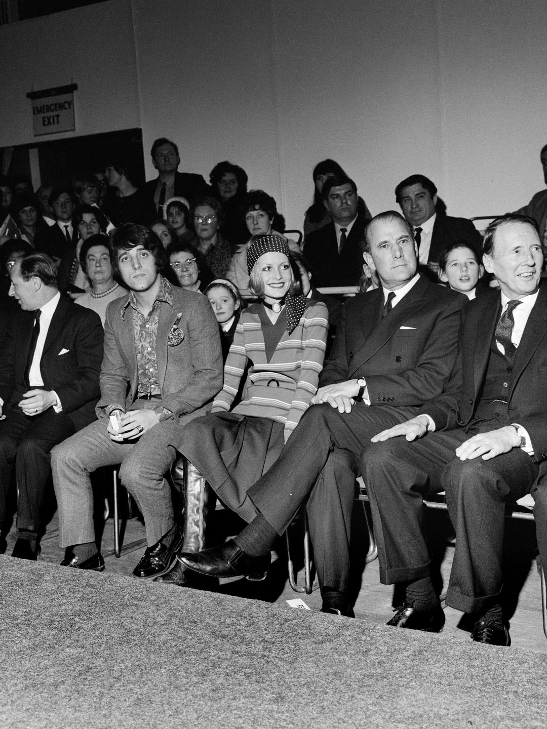 Seated in a front-row seat at a fashion show, Twiggy wears a striped dress and matching headscarf, embodying the vibrant patterns of the 60s. 