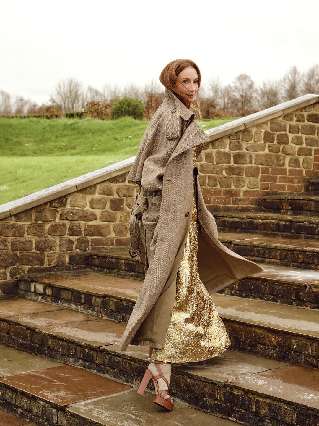 A woman clad in a long tailored coat and metallic gold skirt descends stone steps. Her heeled sandals add an elegant touch to her dynamic stride, set against a lush green background.