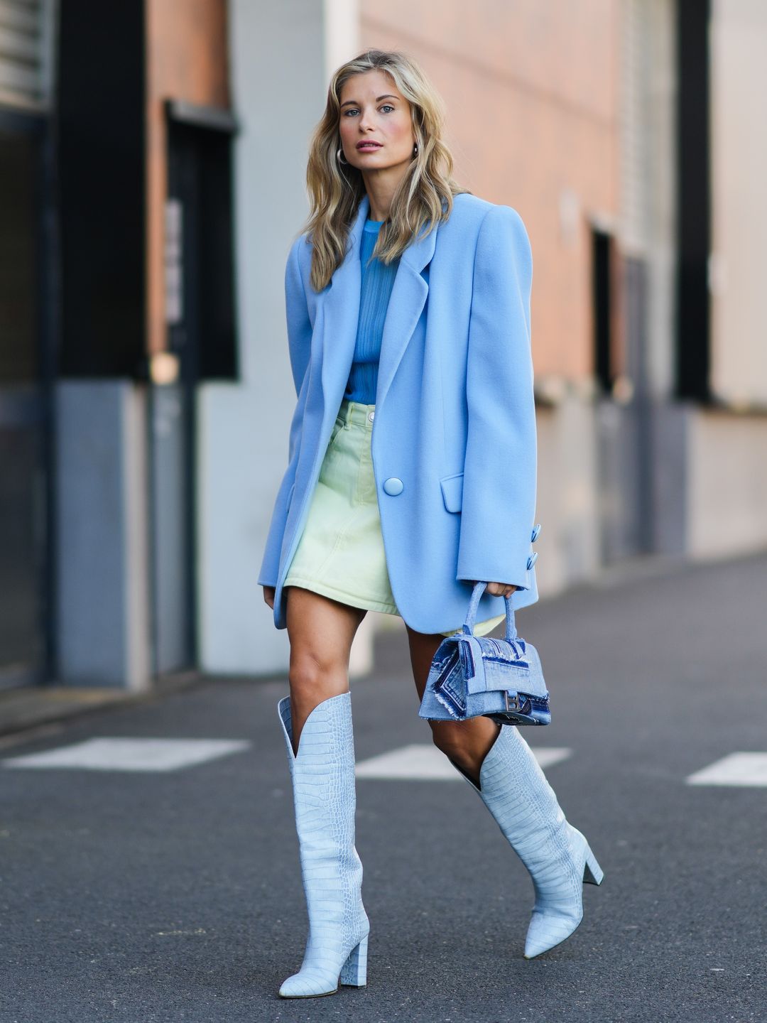 Xenia Adonts wears large silver earrings, a blue ribbed t-shirt, a pale yellow denim short skirt, a pale / baby blue oversized blazer jacket, a blue denim faded ripped Hourglass handbag from Balenciaga, pale blue shiny leather crocodile pattern pointed block heels waders / knees boots, outside Coperni, during Paris Fashion Week - Womenswear Spring Summer 2022
