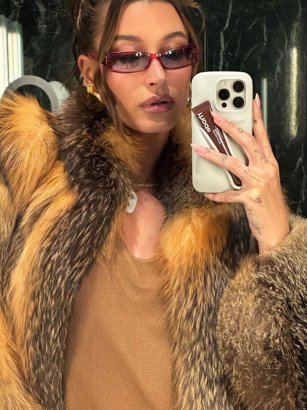 Hailey Bieber wearing a fur coat while taking a mirror selfie with a lip balm-holding phone case