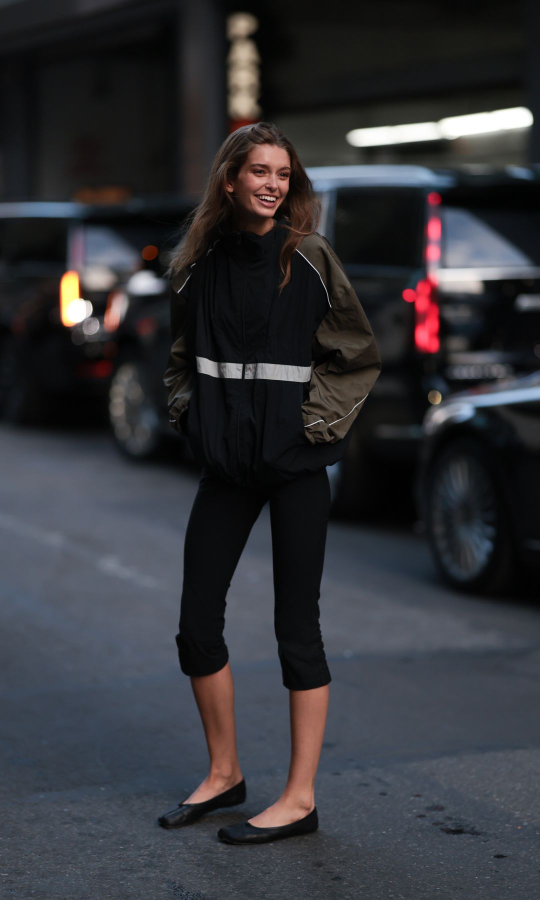 Stella Hanan seen wearing sporty black windbreaker jacket featuring olive-green sleeves, a stand collar, and a reflective white horizontal stripe across the chest, cropped black capri leggings and Maison Margiela black ballet flats, outside the Ralph Lauren Show, during New York Fashion Week