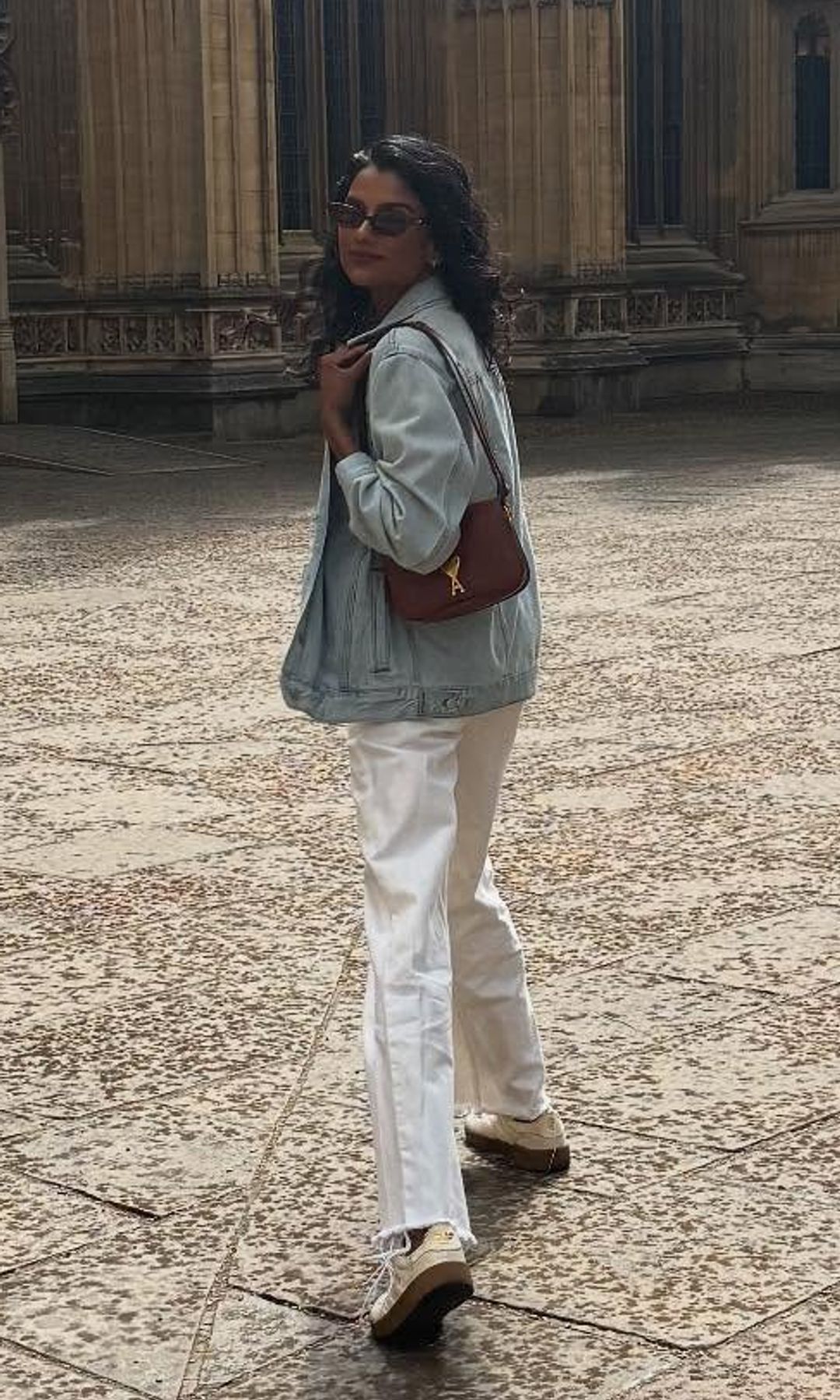 Simone Ashley poses outside of Oxford University in white jeans, sneakers, a black tank top and denim jacket