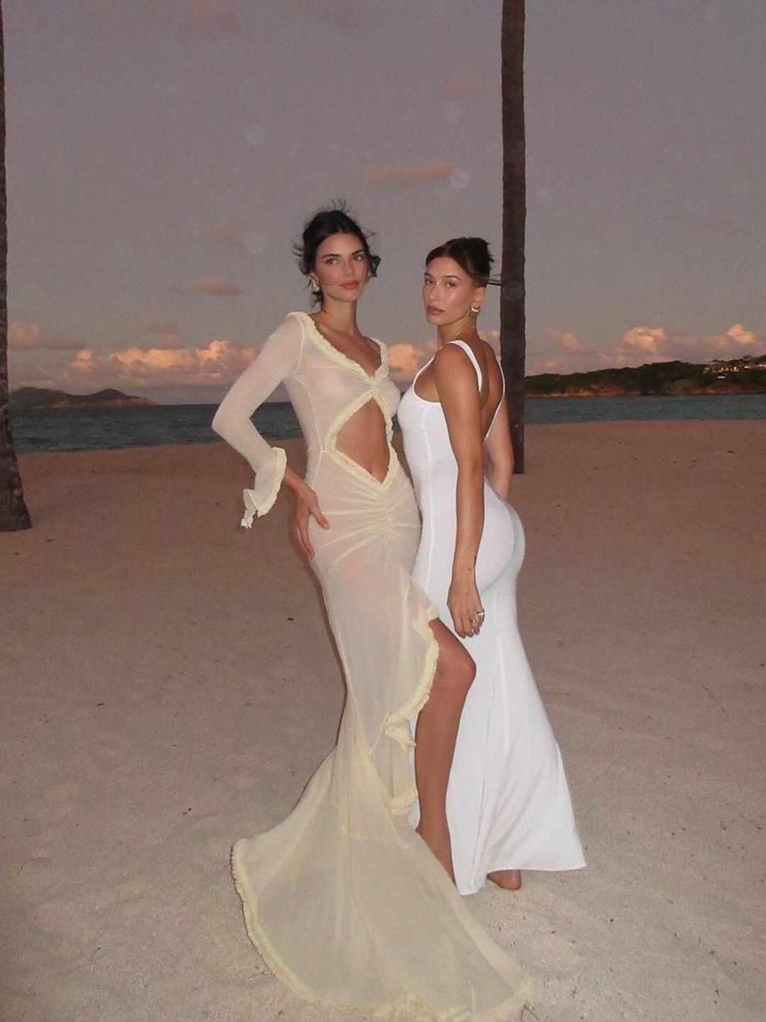 Hailey Bieber and Kendall Jenner pose on a sandy beach in matching cream and white gowns