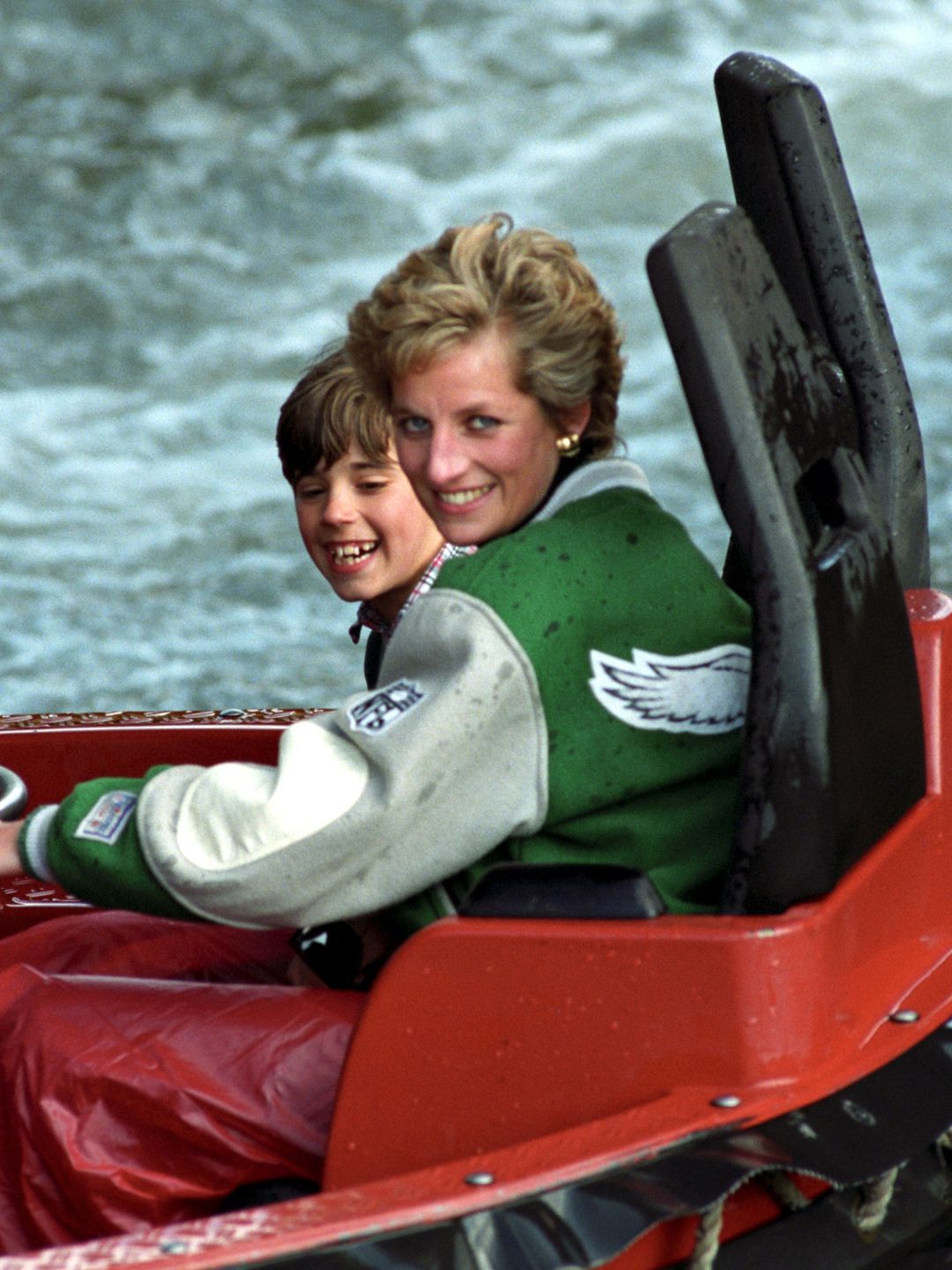 A radiant Princess Diana rides a water raft with young boys, laughing as water splashes around. She wears a green-and-gray Philadelphia Eagles varsity jacket, exuding effortless cool with a motherly touch.