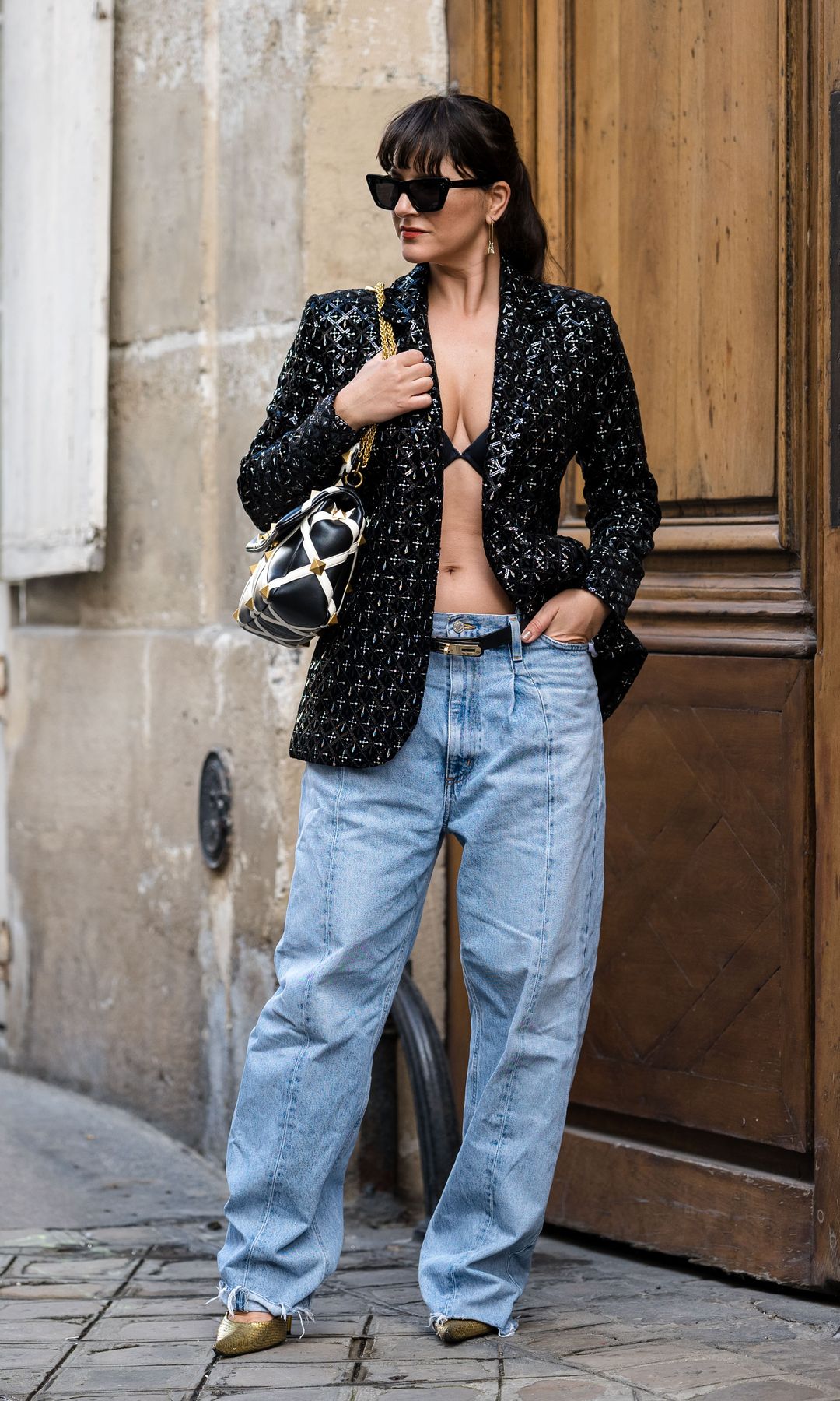 Gili Biegun wears sunglasses, earrings, a black and white studded large bag by Valentino, a black leather belt by Hermes, golden pump shoes by Balenciaga, wide cut low-rise waist jeans / pants by Agold, a black and silver tailored evening blazer jacket by Nadine Merabi, 