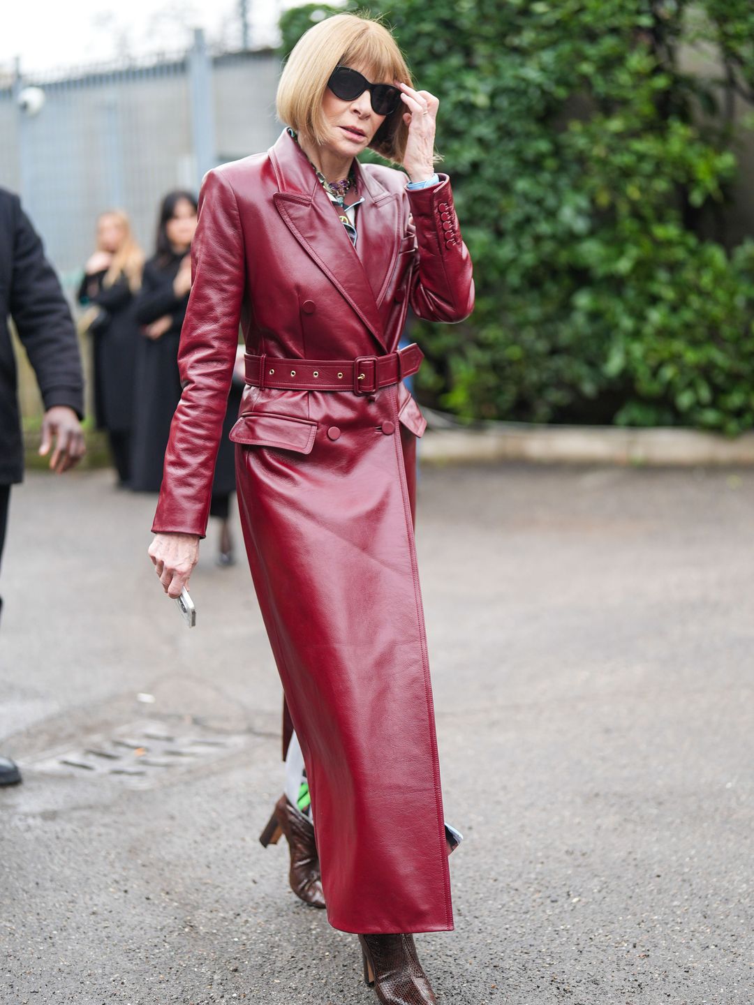 Anna Wintour wears sunglasses, a burgundy leather trench long coat outside Gucci