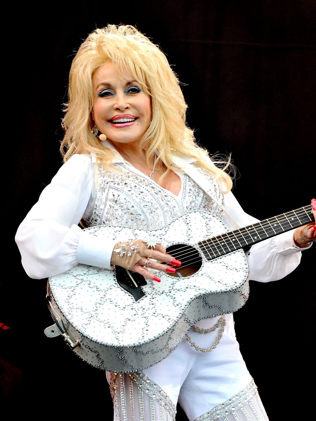 Dolly Parton in a white spangly catsuit while holding a guitar 