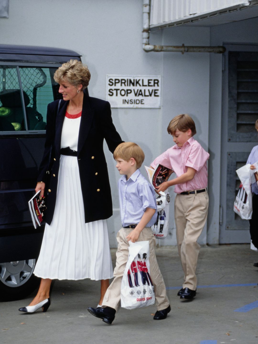 Princess Diana wearing a dark blazer with a pleated white skirt with a young Prince William and Harry  