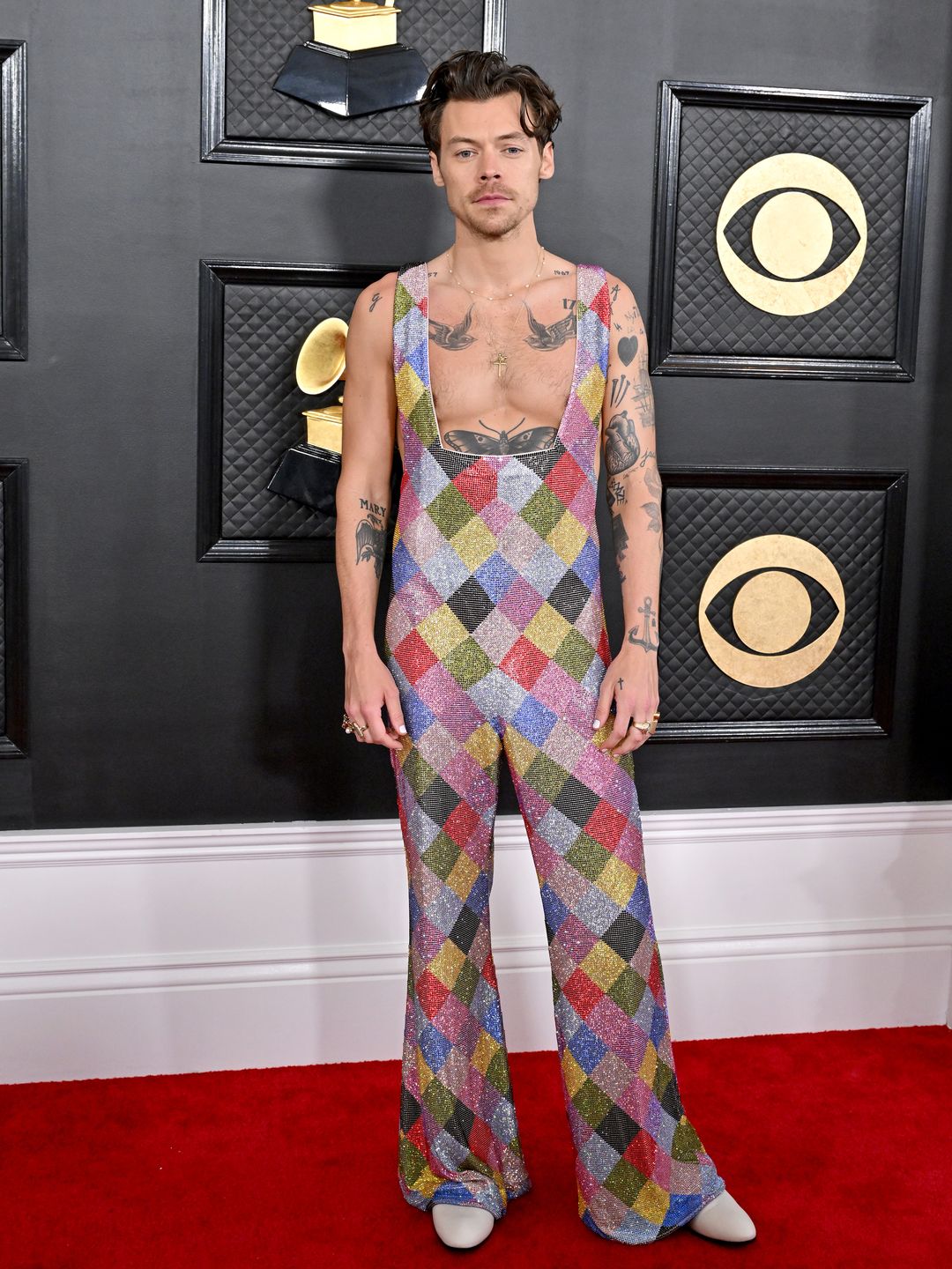 Harry Styles poses on the red carpet at the 2023 Grammy Awards wearing an argyle print multicoloured sequin jumpsuit covered in crystals. It has a low neckline and exposes the swallow tattoos on his chest and his hair is styled with slick curls.