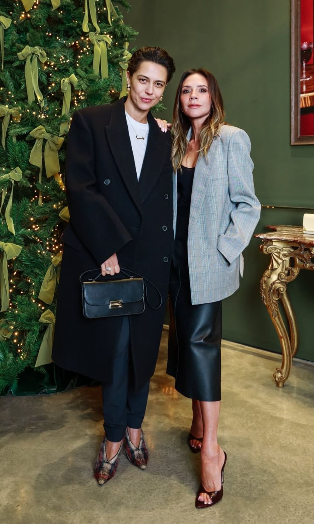 Victoria Beckham poses with a friend in front of a Christmas tree wearing a checkered blazer and leather midi skirt 