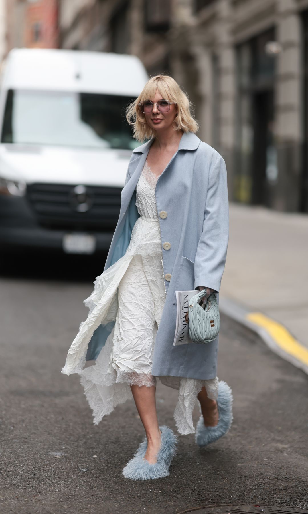 Anna Estrin seen wearing oversized tinted sunglasses with a retro vibe, Daniil Antsiferov white lace dress with crinkled, textured fabric and a scalloped hemline, Natali Zhitko light blue single-breasted coat with oversized buttons and a clean, minimalist cut, draped casually open, Miu Miu soft blue small quilted leather handbag and Akira eye-catching fluffy light blue heels, during New York Fashion Week