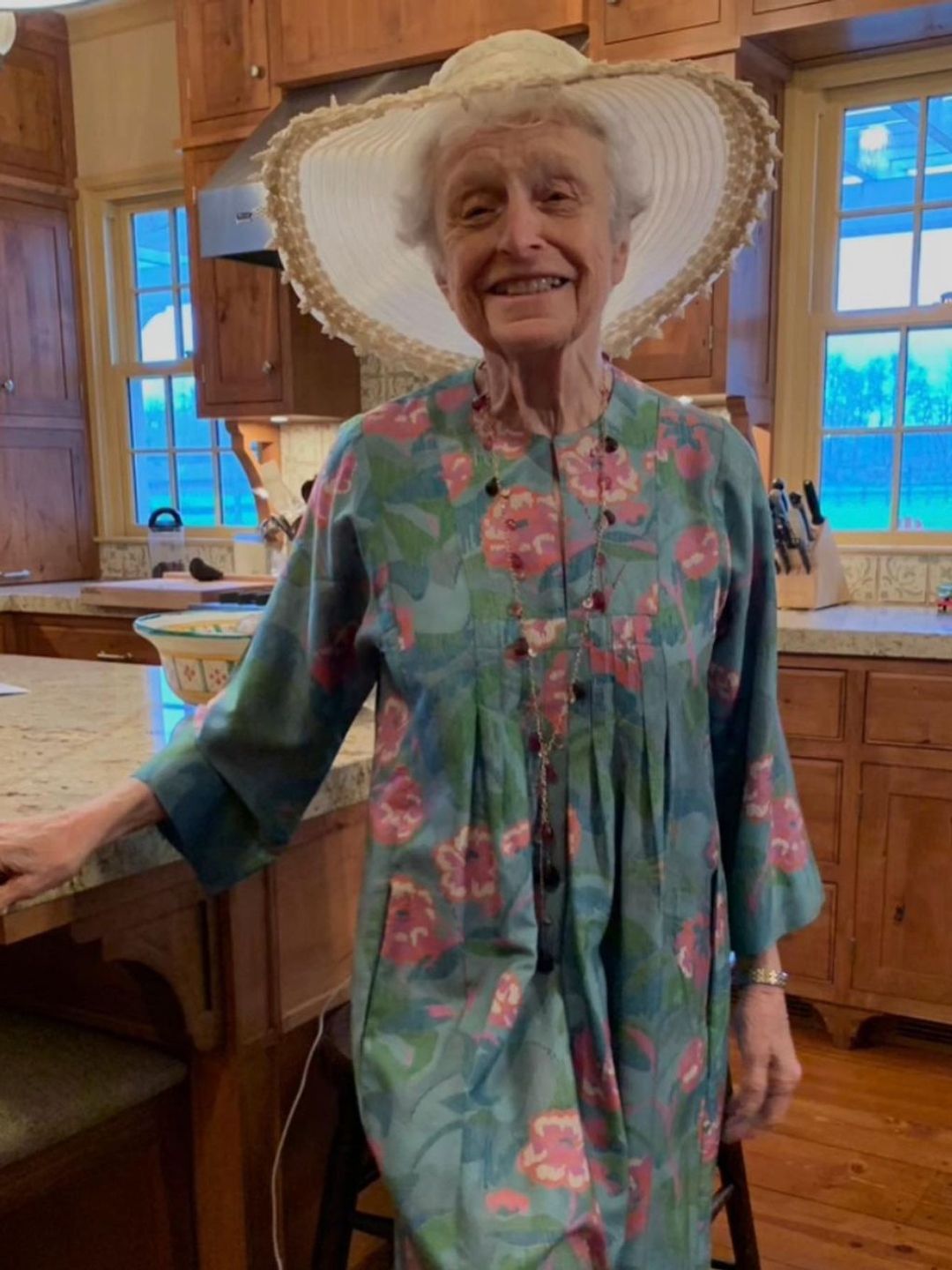 Kyra's mom Patricia smiling in a sunhat and sundress in their kitchen