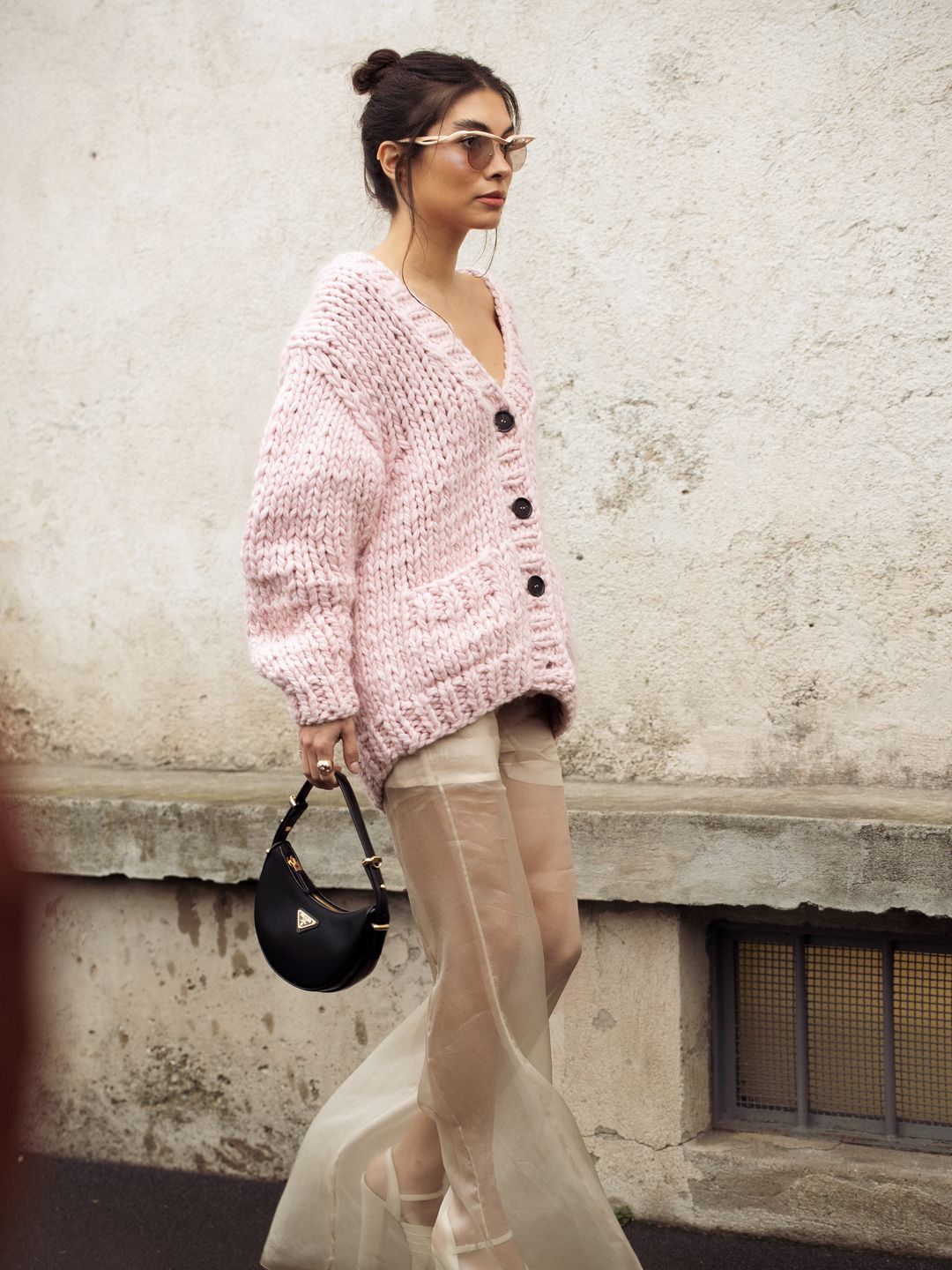 A guest wears a pink knitted cardigan, transparent beige wide pants, white platform shoes, and a black bag outside Prada
