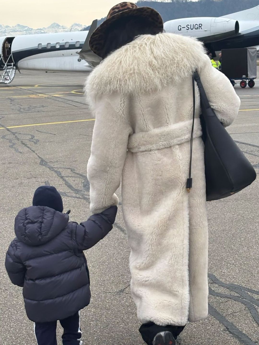 naomi campbell holds hand with her son in front of a private plane, wearing a white shearling coat