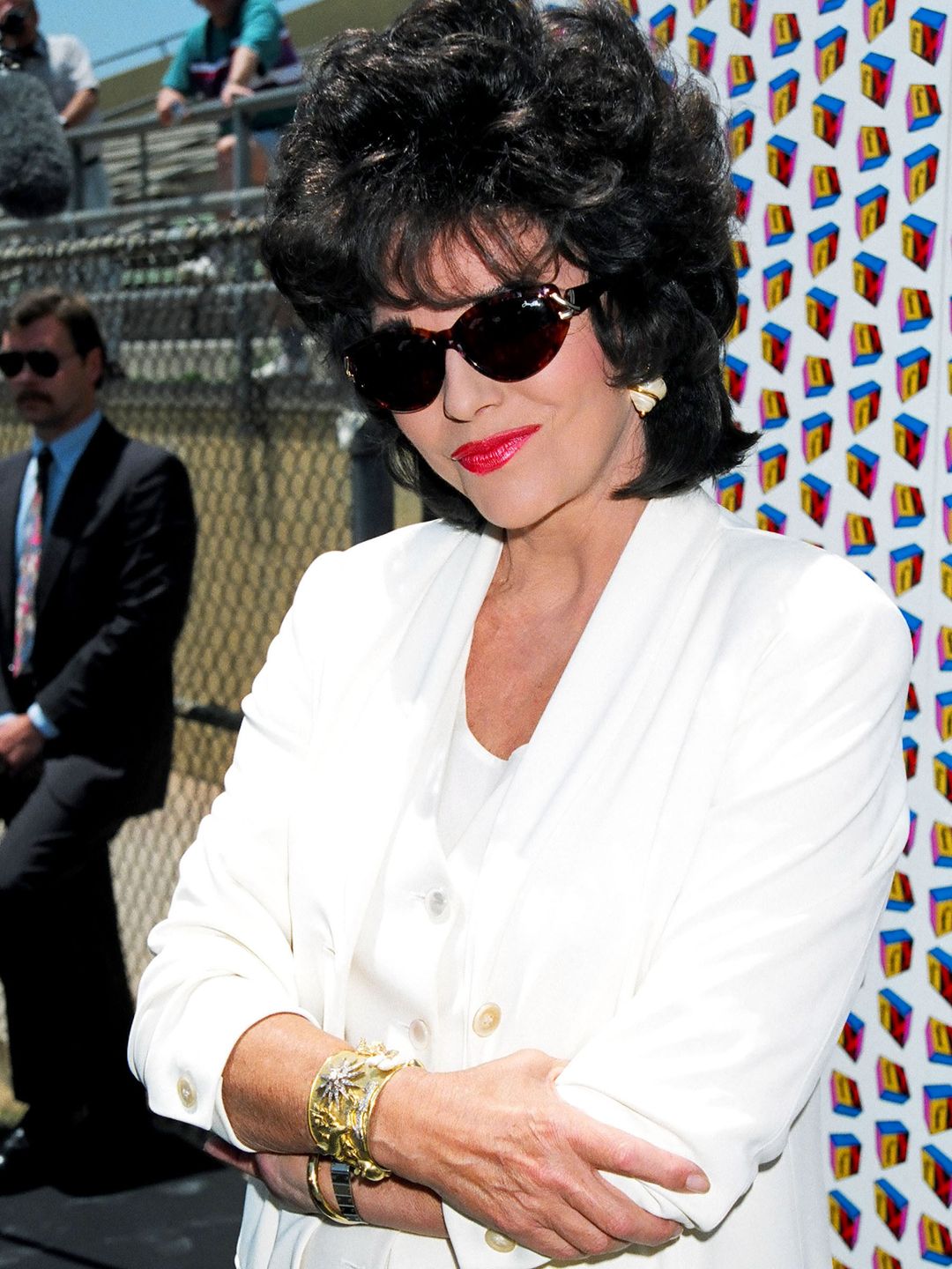 Joan Collins smiling in sunglasses a white suit and red lipstick at an event