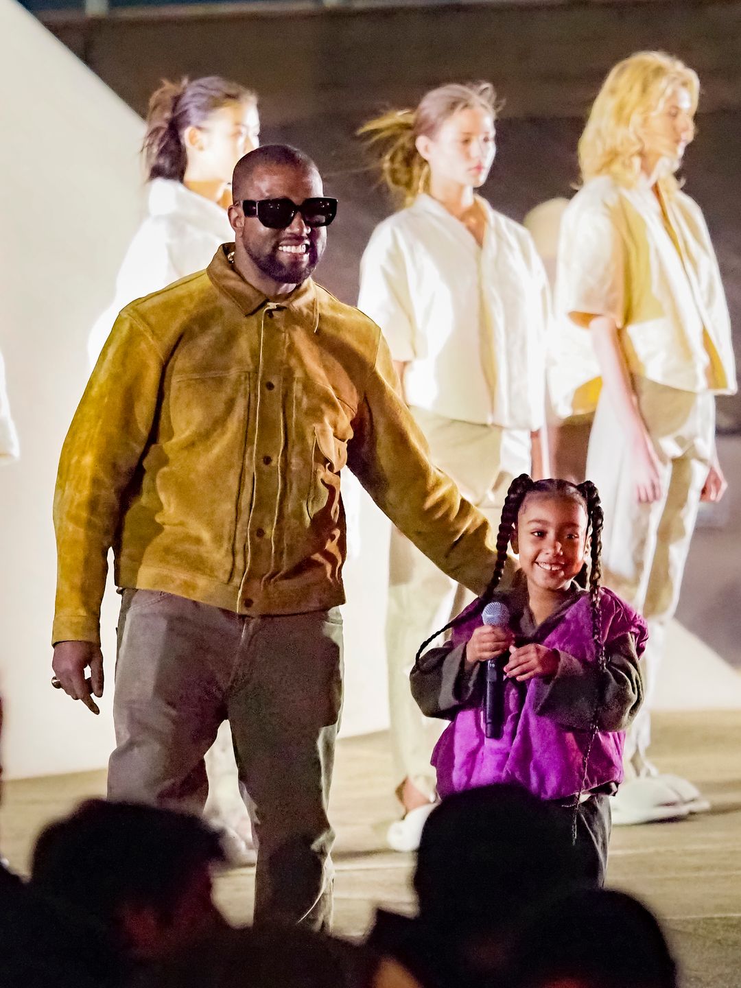 North West smiling on stage holding a mic next to her father Kanye