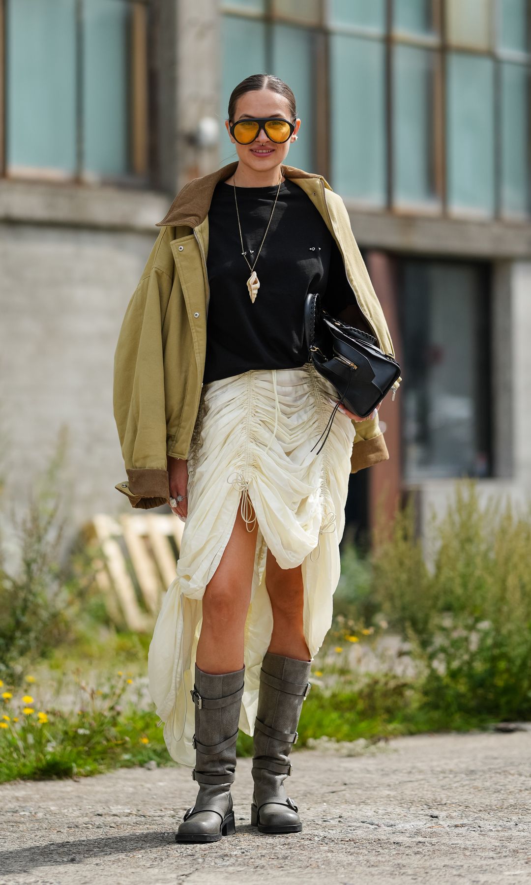 A guest wears dark brown hair styled in a sleek, middle-parted low bun, oversize ambertinted round sunglasses with thin dark frames, and a long goldtone chain necklace with a cream pendant; an oversized khaki canvas jacket with a brown collar and visible brown lining is draped over the shoulders above a black crewneck Tshirt; an ivory ruched drawstring skirt in lightweight semisheer fabric with an asymmetric draped hem is paired with a small structured black patentleather tophandle bag with silvertone hardware; gray leather midcalf boots with round toes, low block heels, and strapandbuckle details complete the look; soft neutral lipstick is visible; posing, outside Marimekko, during Copenhagen Fashion Week
