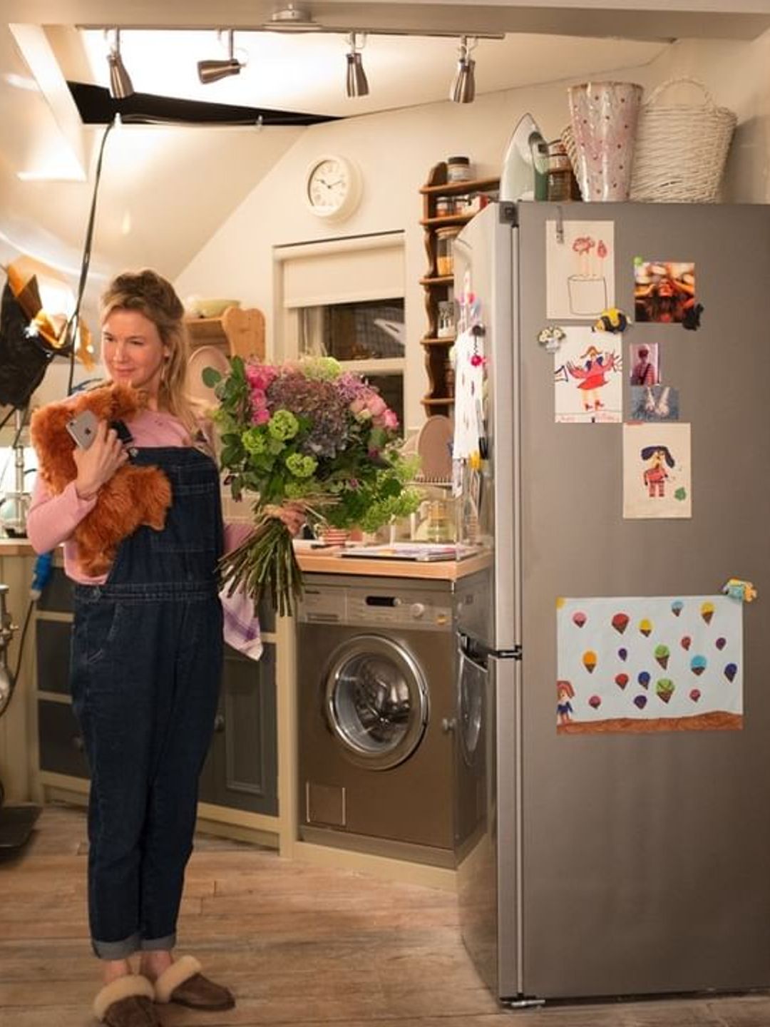 Bridget Jones stands in her kitchen holding flowers and wearing denim overalls 