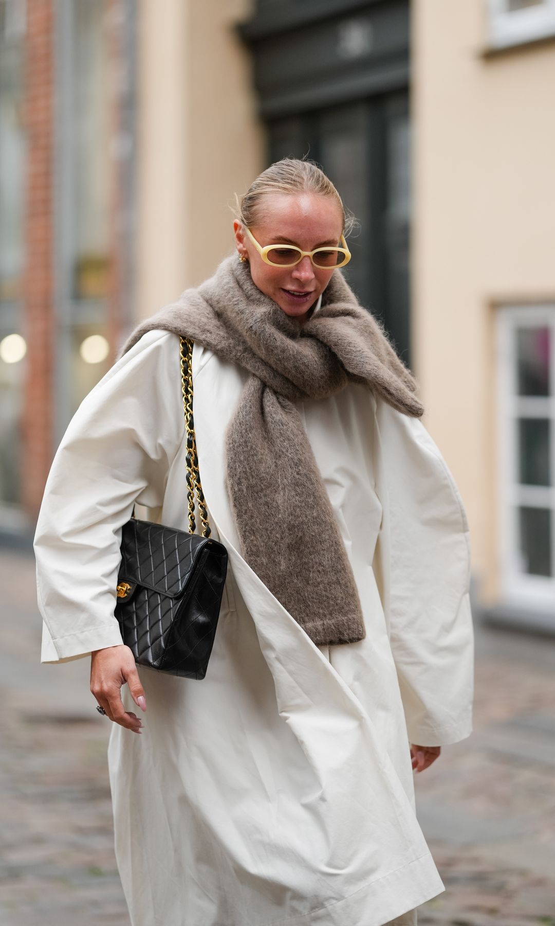 A guest wears blonde hair slicked back into a low bun, small oval sunglasses with pale yellow acetate frames and dark lenses, a chunky taupe brushed-knit scarf wrapped around the neck and draped over the shoulders, an oversized off-white coat with dropped shoulders and wide sleeves, and a black quilted leather flap shoulder bag with a gold-tone chain-and-leather strap and a gold CC turn-lock, with nude glossy nail polish, walking, outside Nicklas Skovgaard, during Copenhagen Fashion Week