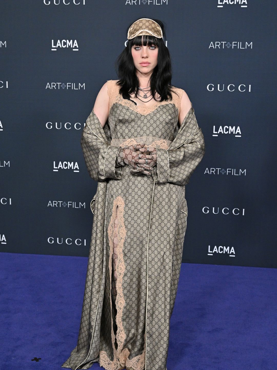  Billie stuns in a Gucci logo olive-green satin gown with lace trim. She accessorizes with a matching sleep mask on her head, soft black hair, and layered necklaces.