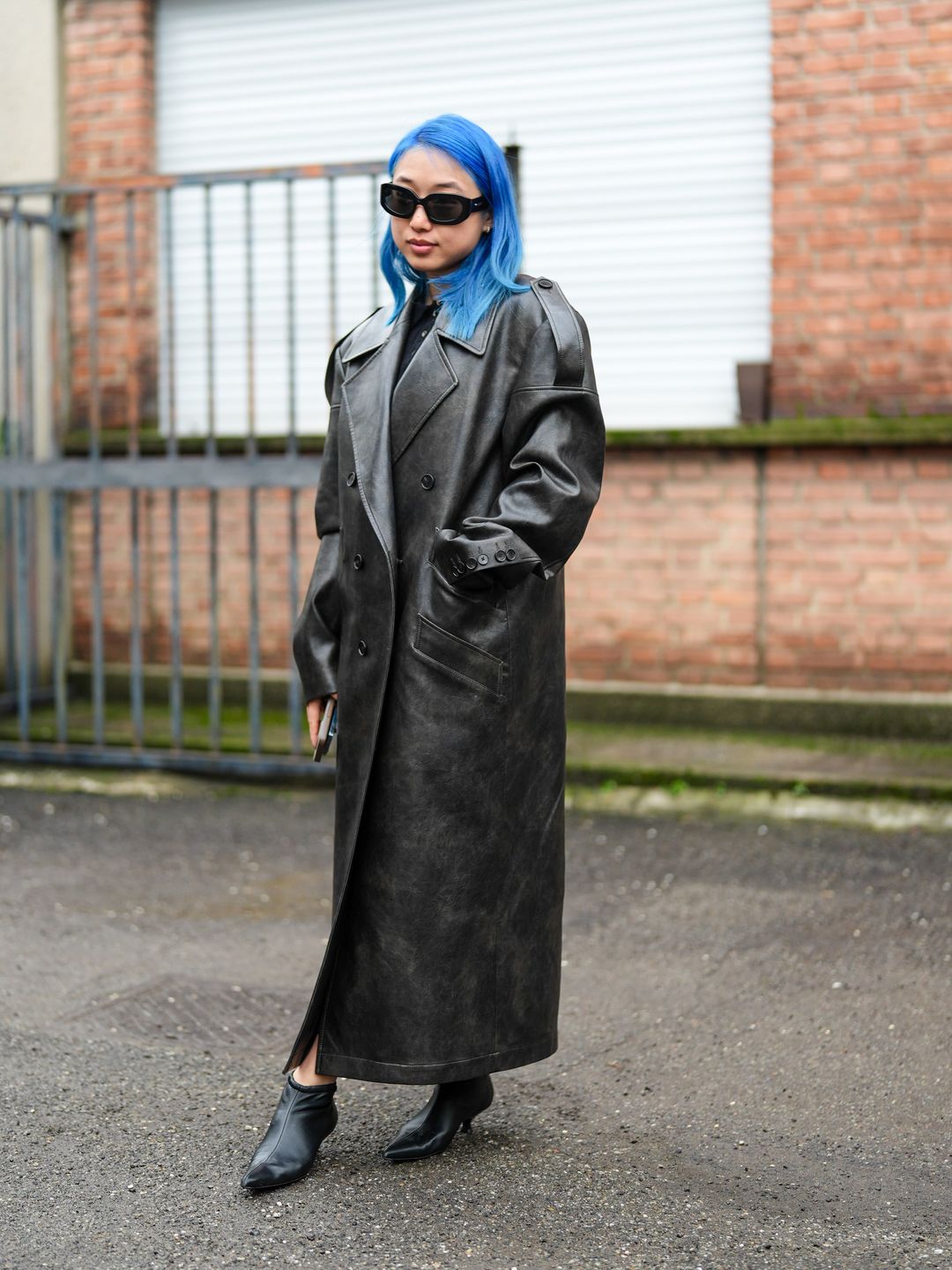 Margaret Zhang wears sunglasses, a black leather double breasted trench long coat, kitten heels black leather pointed shoes outside Gucci