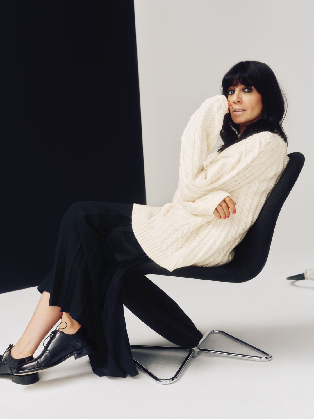 Claudia Winkleman siedzi na krześle, ubrana w kremowy sweter i czarny kilt 