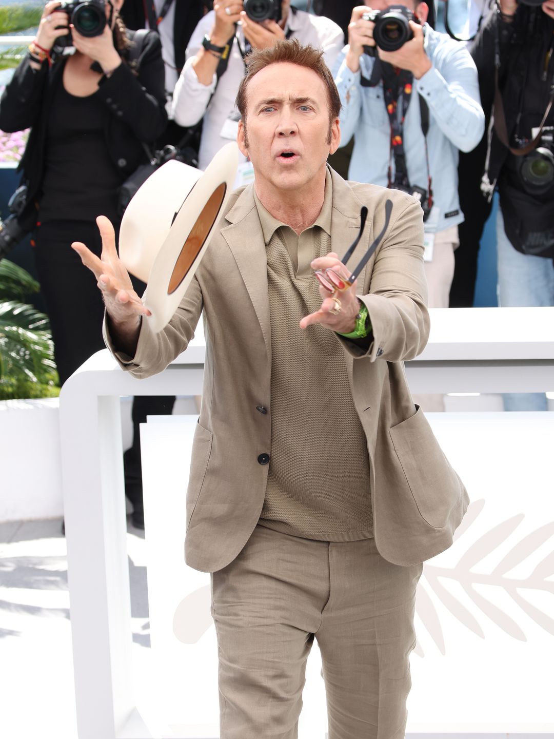 Nicolas Cage in a beige outfit trying to catch a hat