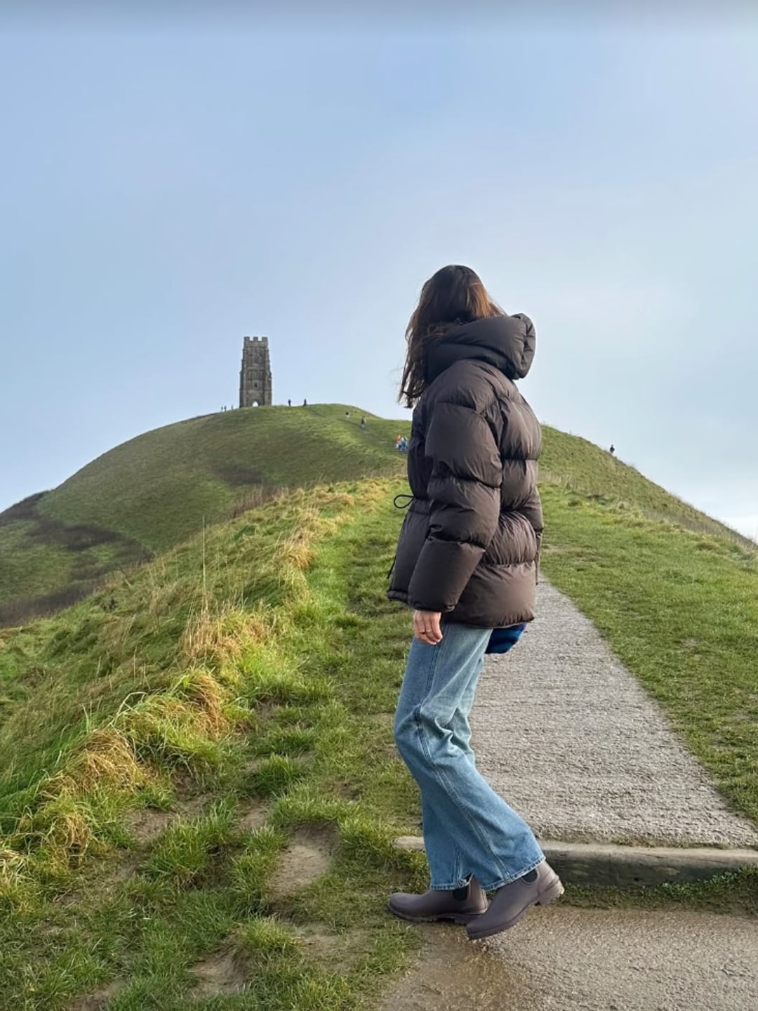 Alexa Chung wears a brown puffer coat, straight-leg jeans, and boots while strolling near Glastonbury Tor—practical and stylish.