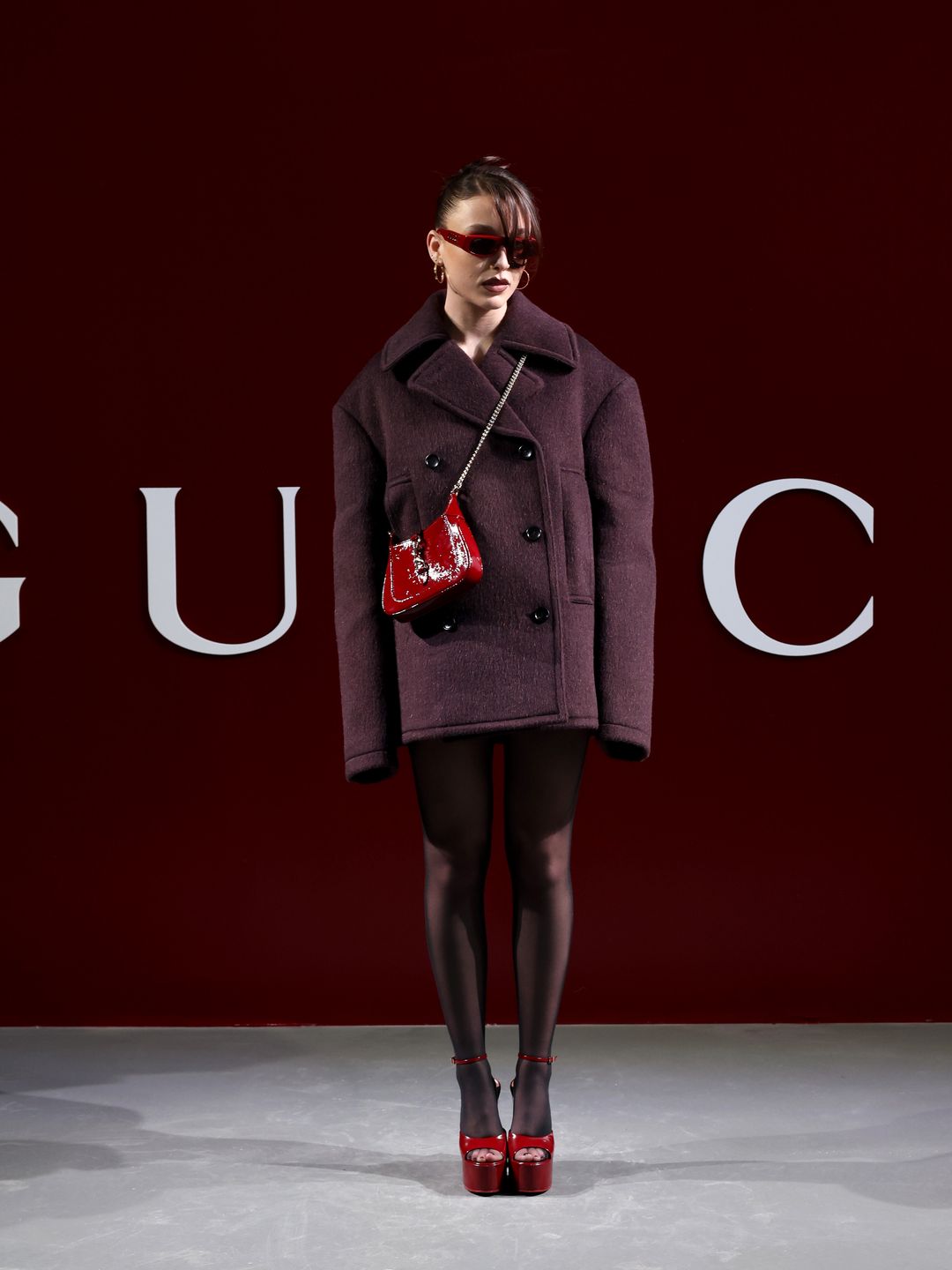 Emma Chamberlain attends the Gucci Women's Fall Winter 2024 Fashion Show during Milan Fashion Week  in a burgundy coat, sheer tights, red shoes and a red bag