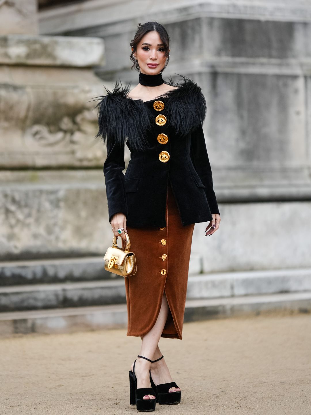 Heart Evangelista wears a chiker, a black jacket with fluffy shoulder pads and large golden buttons, a brown lustrous silky slit midi skirt, platform high heels shoes, outside Schiaparelli, during the Haute Couture Spring/Summer 2024