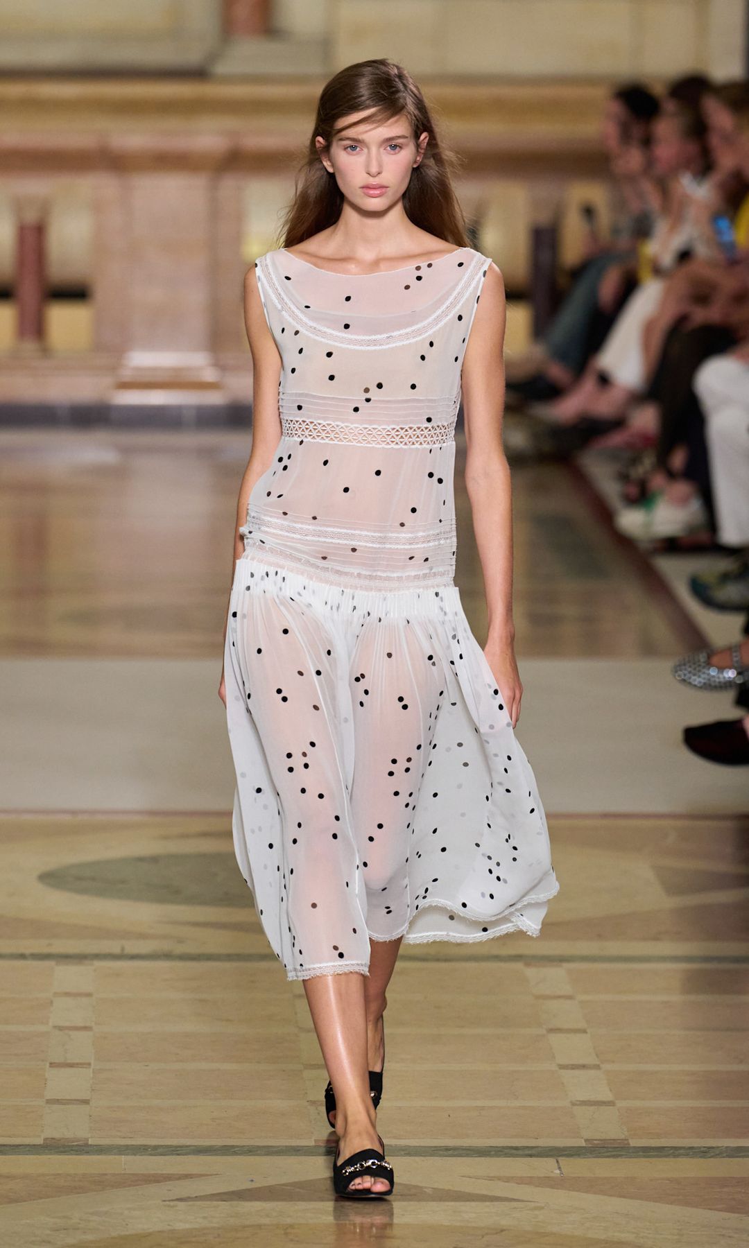 A model walks the runway at Tory Burch in a sheer white dress with drop waist, covered in black polka dots teamed with black sandals