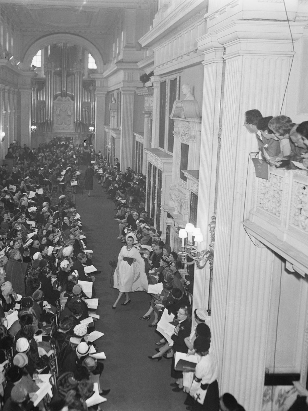 This vintage black-and-white photograph captures a mid-20th century fashion show at Blenheim Palace. Models walk down a grand, column-lined hall while elegantly dressed attendees sit closely on either side of the runway. The elaborate setting, with its high ceilings and ornate architecture, adds a regal air to the event.