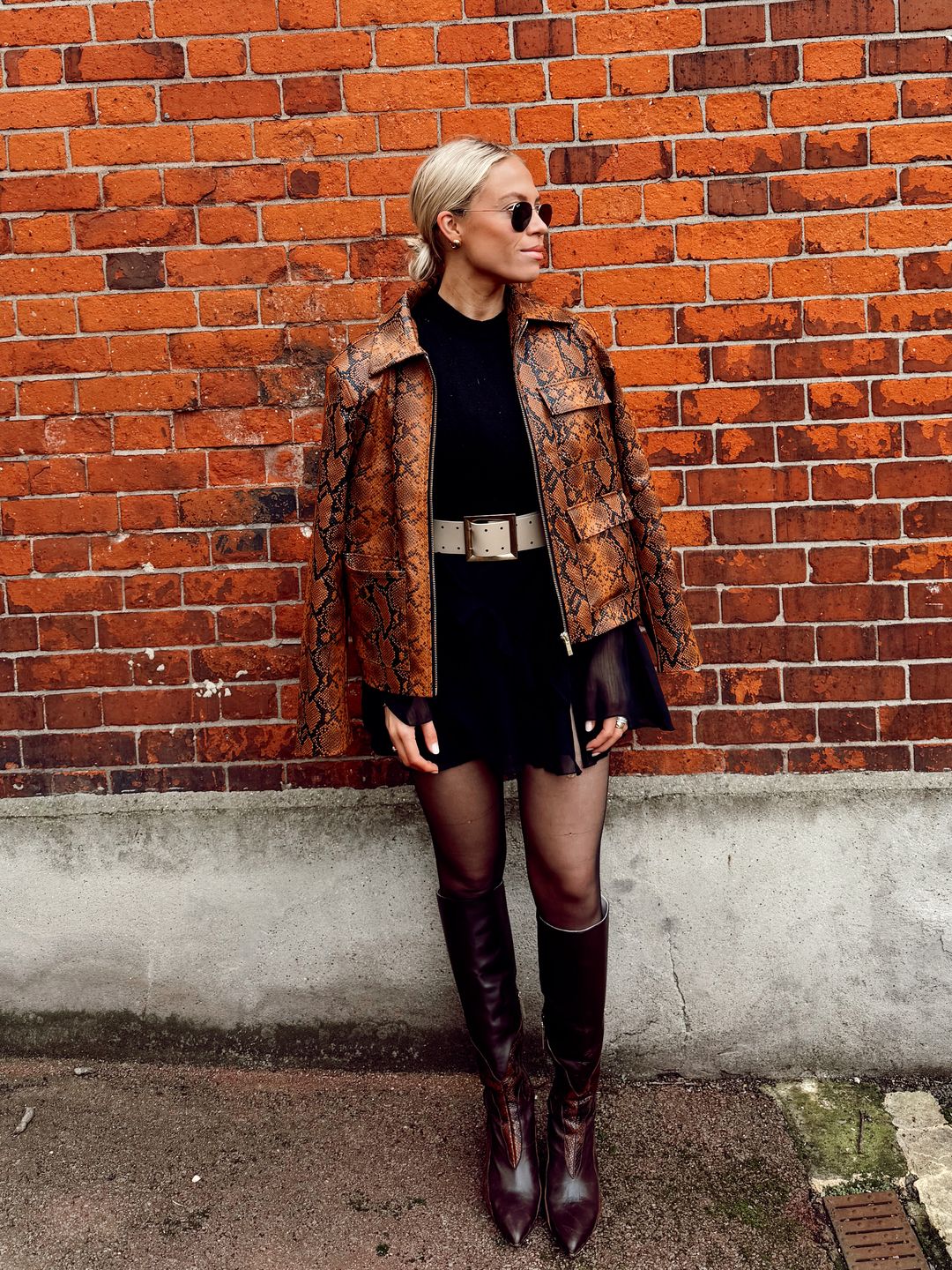 Lauren Ramsay poses in front of a brick wall at fashion week in a pair of knee high boots, a brown jacket and sunglasses.