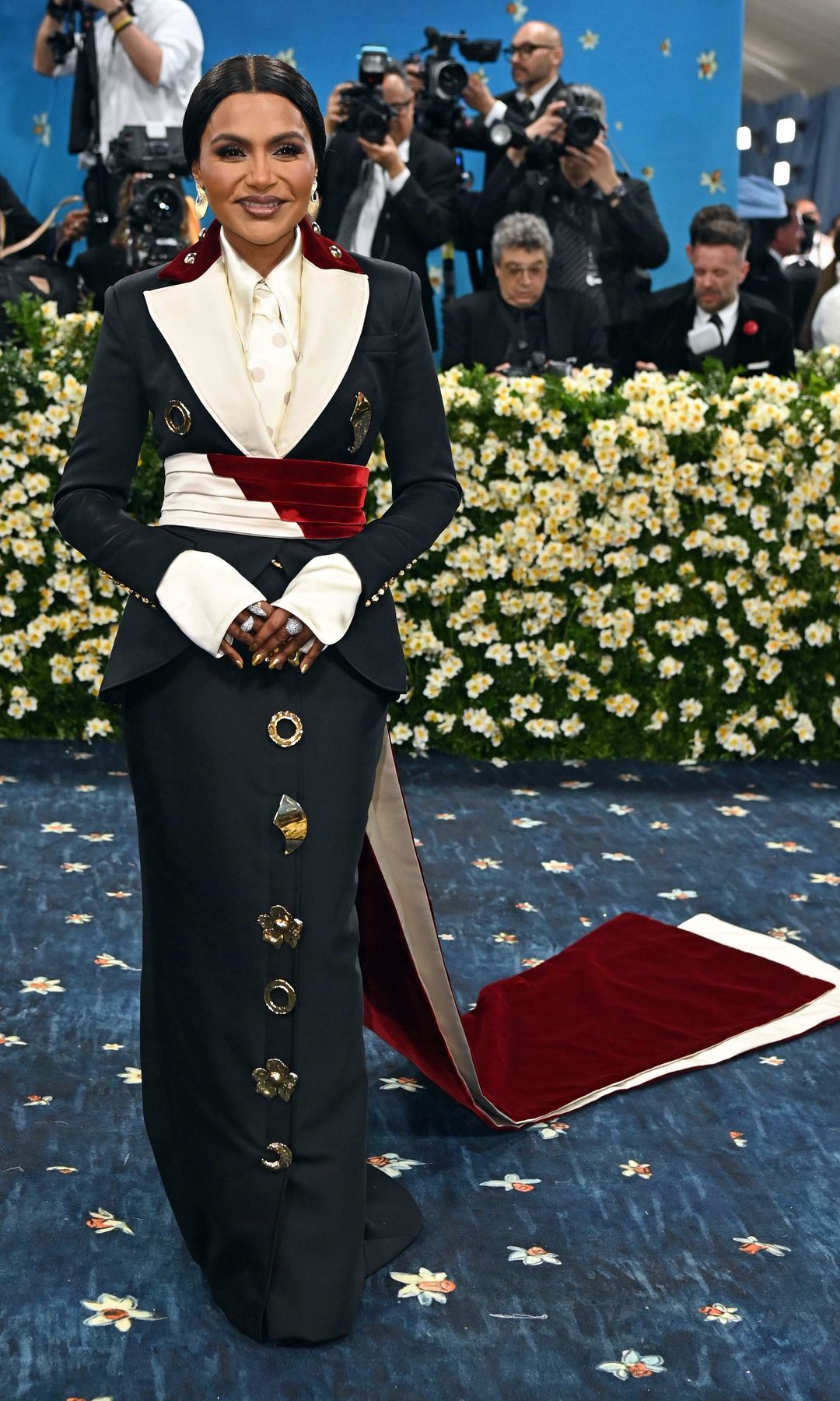 Mindy Kaling wears a black skirt and blazer, red and gold train, white shirt and cream tie paired with big gold earrings.