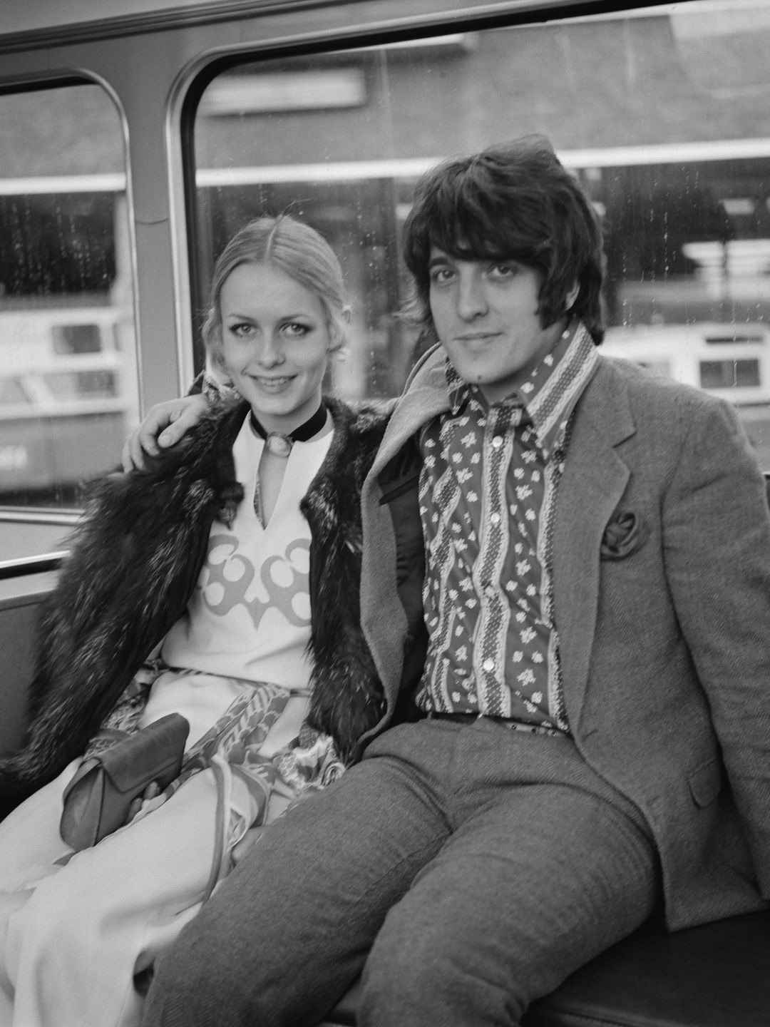 Relaxed on public transport, Twiggy looks effortlessly stylish in a fur-collared coat paired with a printed dress. Her bohemian-inspired outfit contrasts with the structured interior of the bus