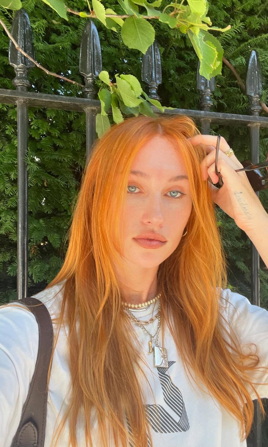 Model Miki Davis looks to the camera in front of a leafy green setting. Her hair is long, vibrant orange with a middle part and gentle layers. 